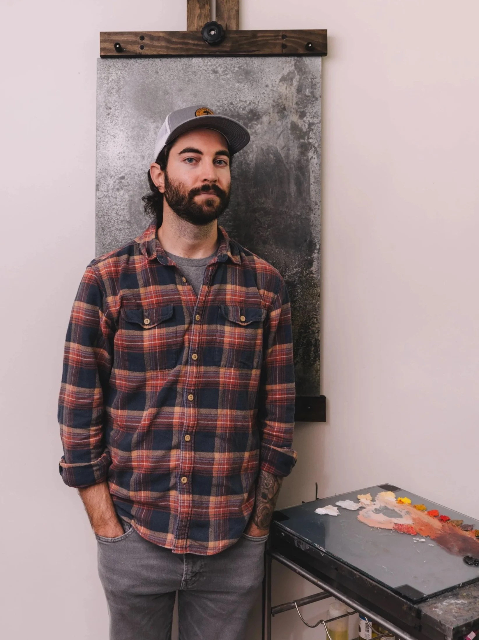 A man with a beard and tattoos on his left arm, wearing a gray cap and an orange plaid shirt, stands next to an art studio table with mixed paint colors. Behind him is an abstract textured painting on a wall, hanging from a wooden frame.