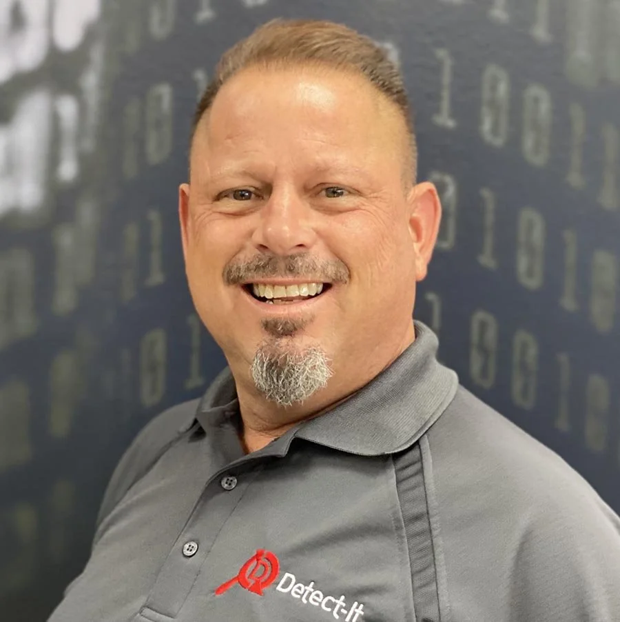 Headshot of a smiling man wearing a gray polo shirt with a red logo and the words 'Detect-it'. background is blurred with numbers or codes.
