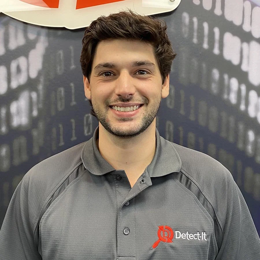 A smiling young man with dark brown hair wearing a gray polo shirt with a Detect It logo on the chest, standing in front of a dark background with a pattern of binary code.