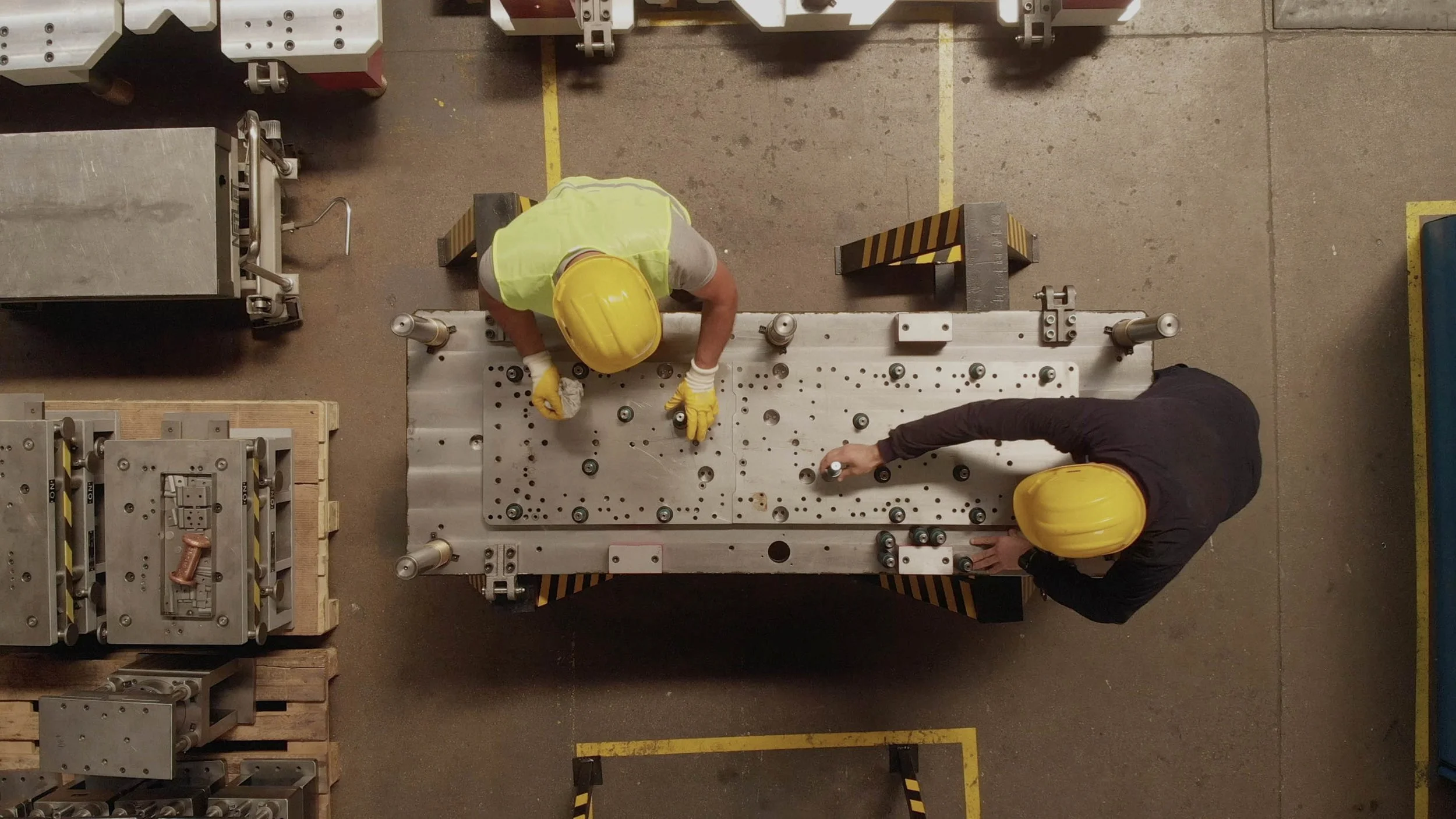 Two workers wearing yellow safety helmets and gloves working on a large metal component in a factory or industrial setting, surrounded by various machinery and equipment.