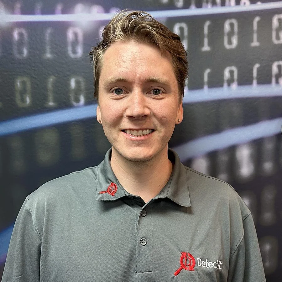 A young man with short brown hair smiling, wearing a gray polo shirt with a red embroidered logo and name tag, standing in front of a blurred digital data background.