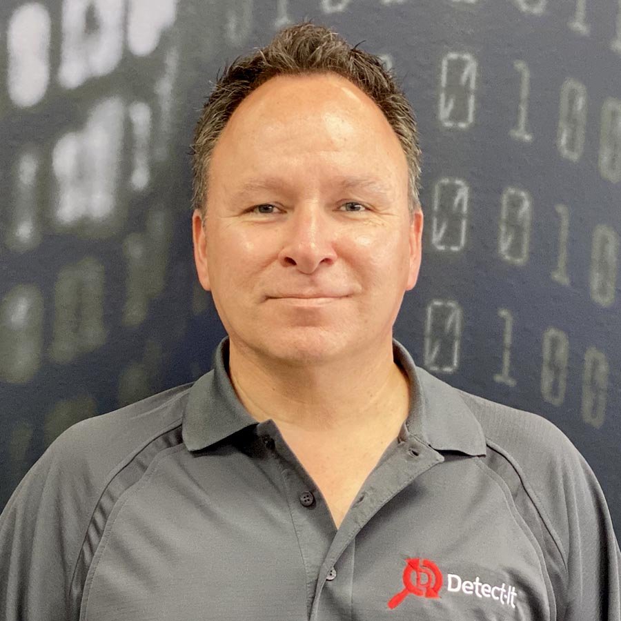 A man wearing a gray collared shirt with a 'DetectIt' logo standing in front of a dark background with binary code.