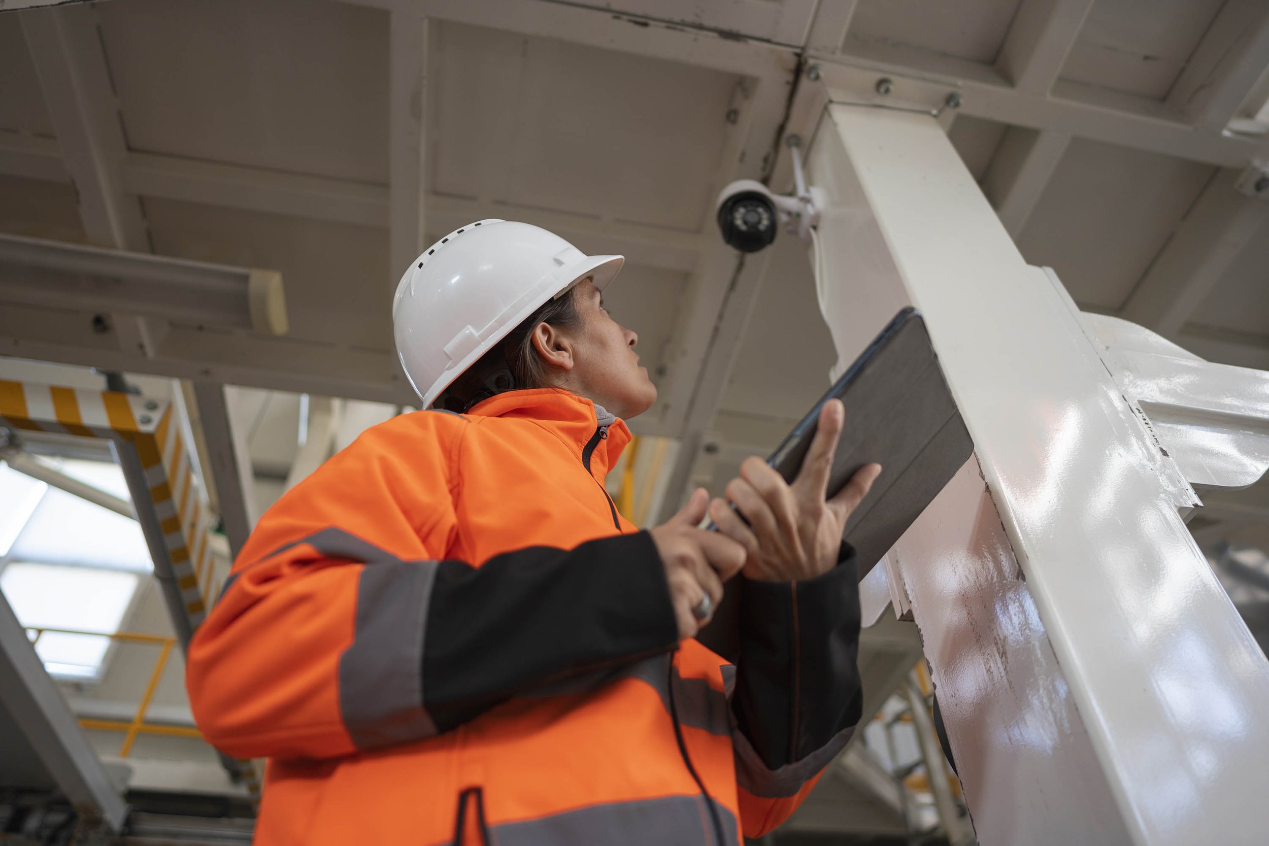 A worker wearing an orange safety jacket and white safety helmet inspecting or working on a large industrial structure or equipment using a tablet inside a factory or industrial facility.
