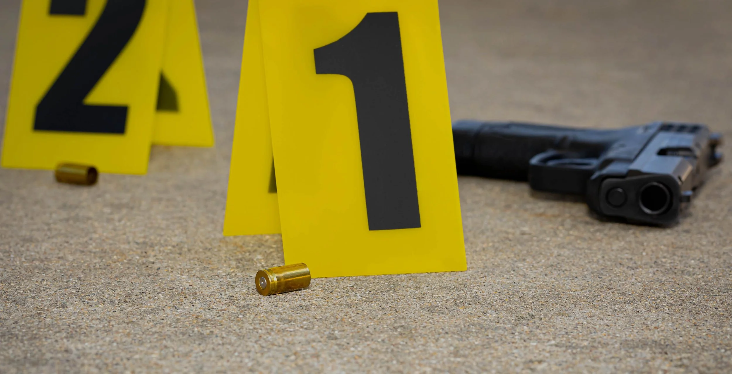 Yellow evidence markers numbered 2 and 1, black handgun lying on a surface, and spent bullet casings, indicating a crime scene investigation.