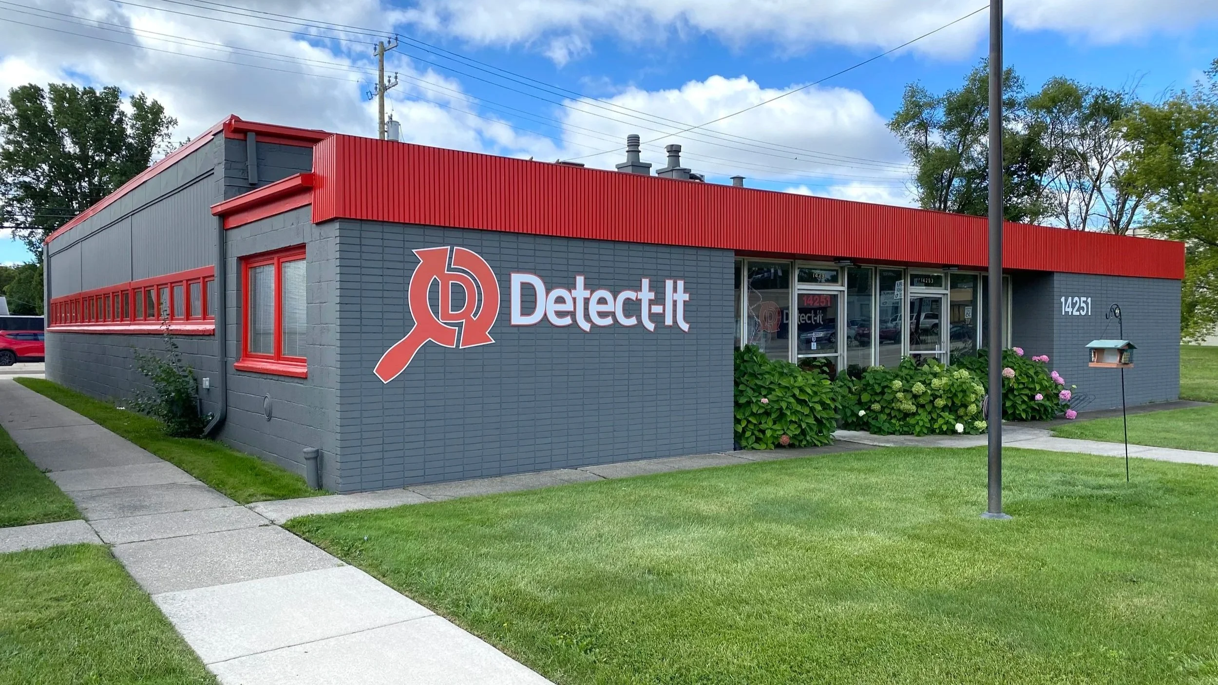 A gray building with red trim and a sign that reads 'Detect-it' in white letters with a magnifying glass logo. The building has large windows and is surrounded by green grass and shrubbery, with a sidewalk in front. There are blue skies with some clouds and trees in the background.