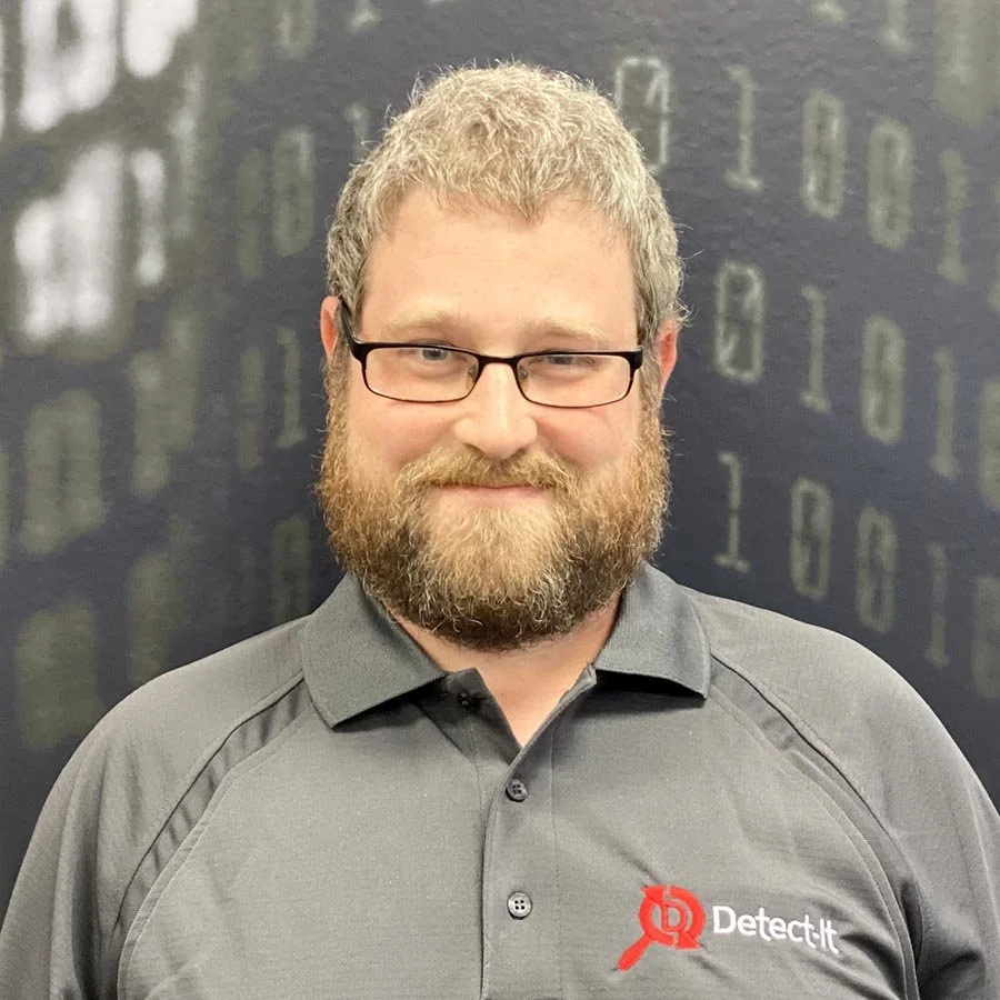 A man with glasses, a beard, and curly hair wearing a gray polo shirt with a Detect it logo standing in front of a dark background with binary code.