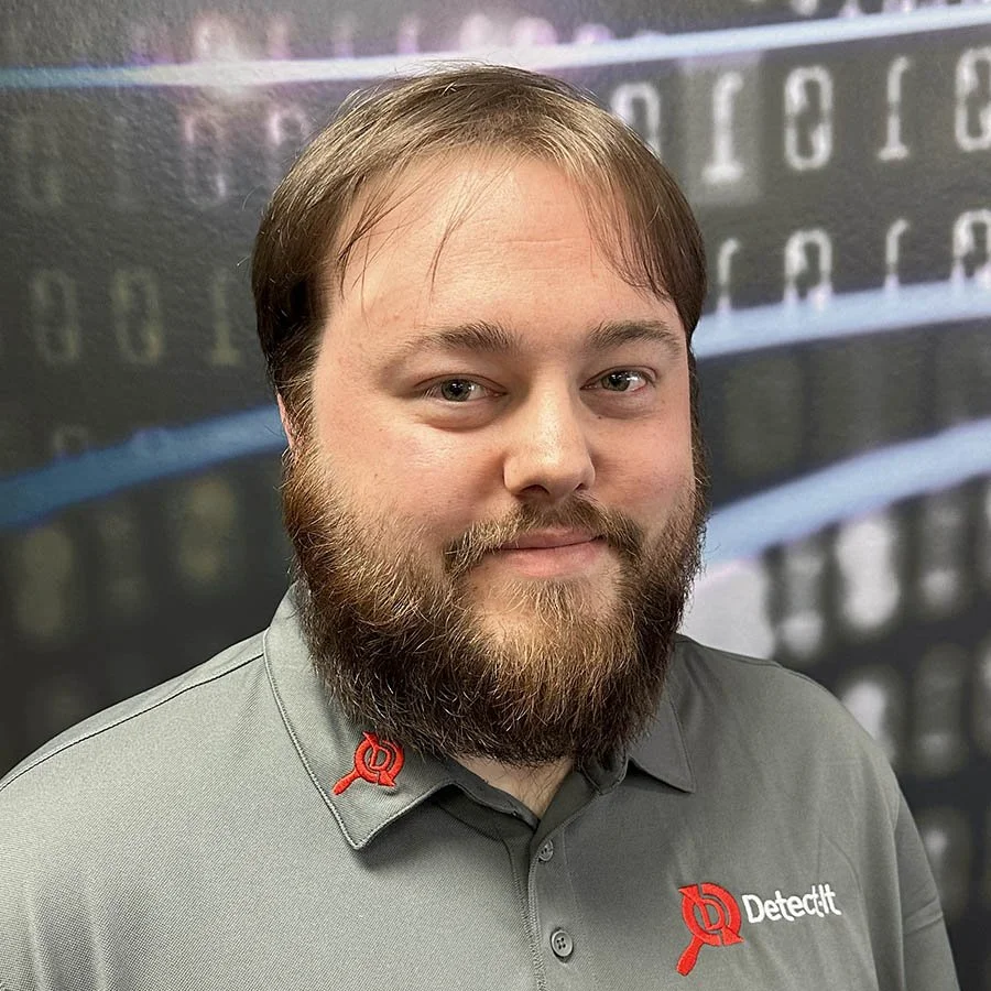 Man with a beard and short brown hair wearing a gray polo shirt with a red logo and the word 'Detectit' on it, standing in front of a background with binary code.