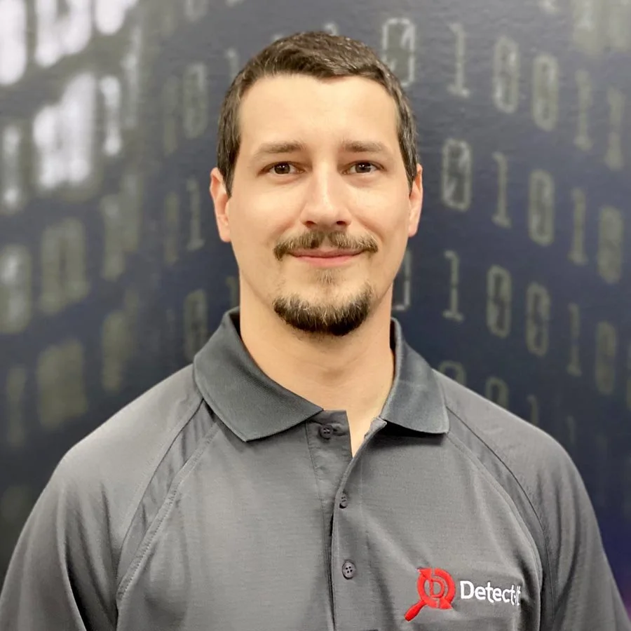 A man with short dark hair, a beard, wearing a gray branded polo shirt, standing in front of a dark background with faint binary code.