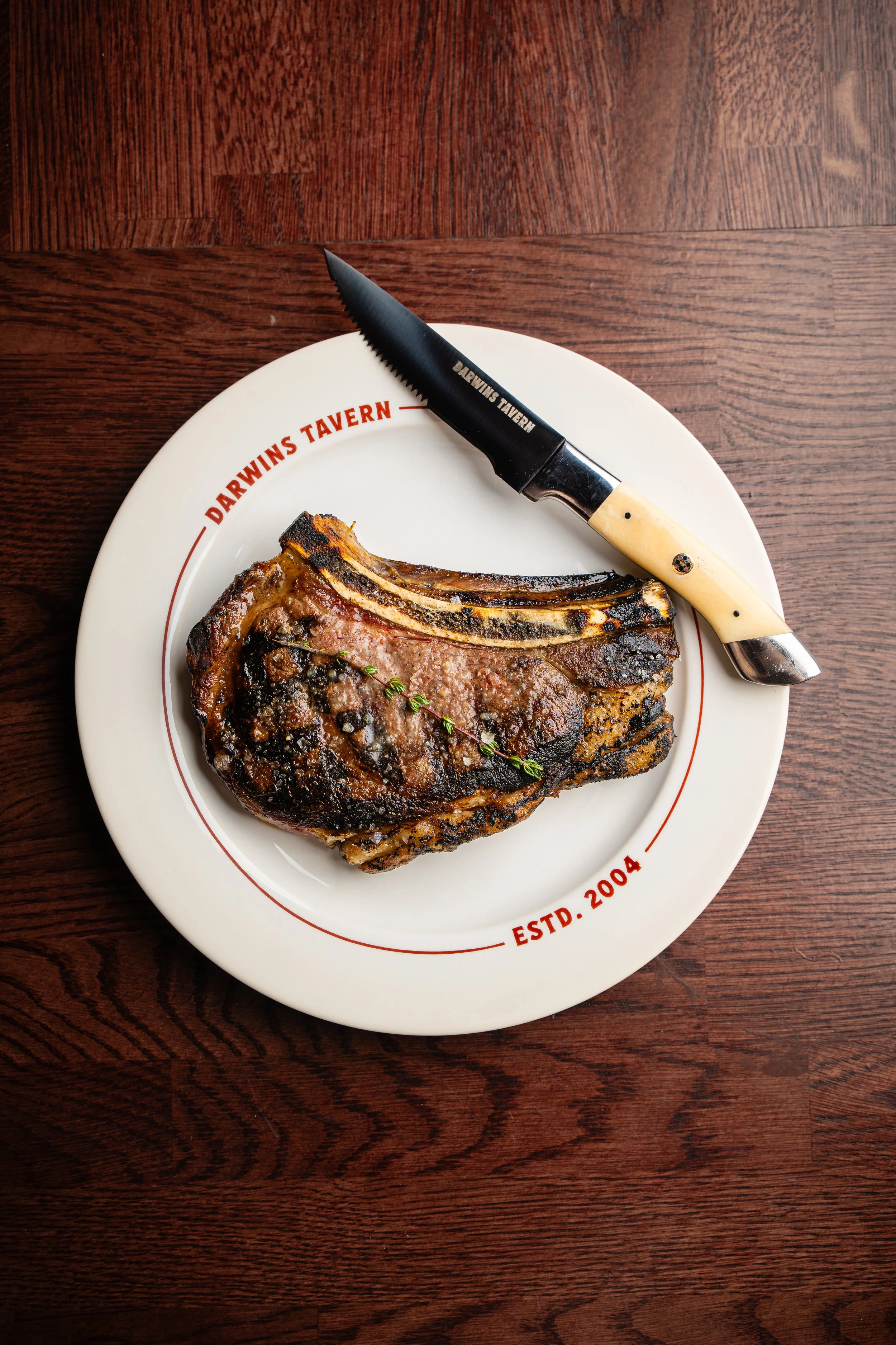 A plate with a cooked steak garnished with a sprig of thyme and a knife on top, on a wooden table. The plate has red lettering that reads "Darwins Tavern" and