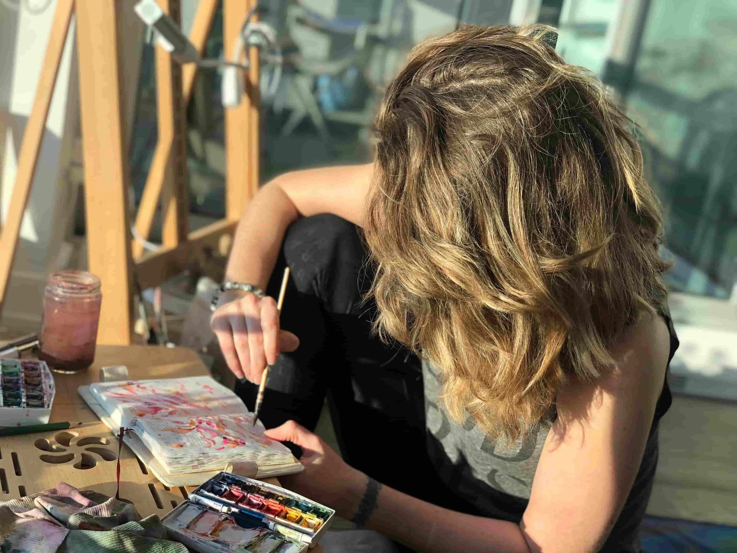 early morning light showing a woman artist with wavy blond hair doing a watercolor painting in her small journal in a studio surrounded by large windows