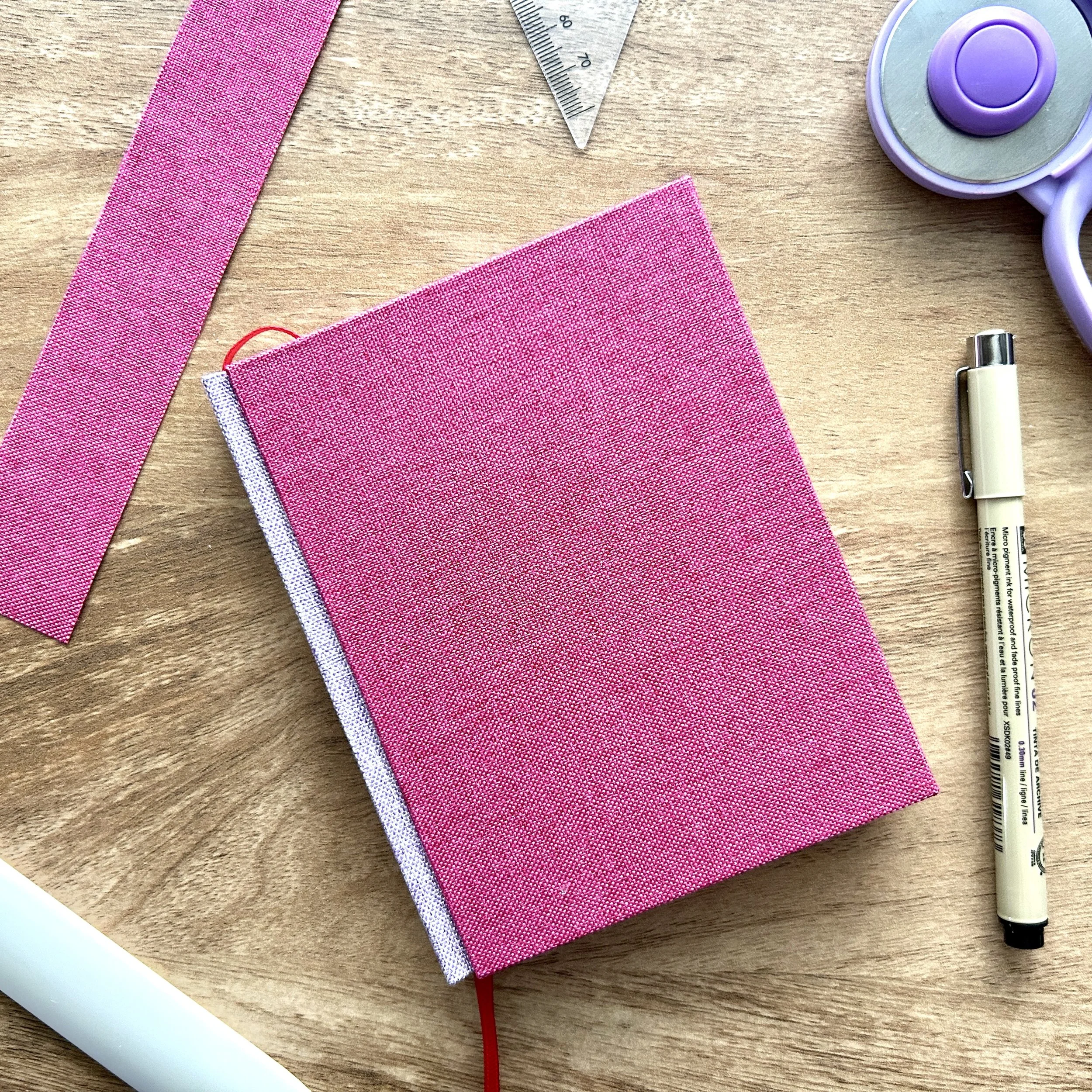 Various sewing and craft supplies arranged on a wooden surface, including pink fabric strips, a silver ruler, a pink fabric square, a white pen, and a purple tape dispenser.