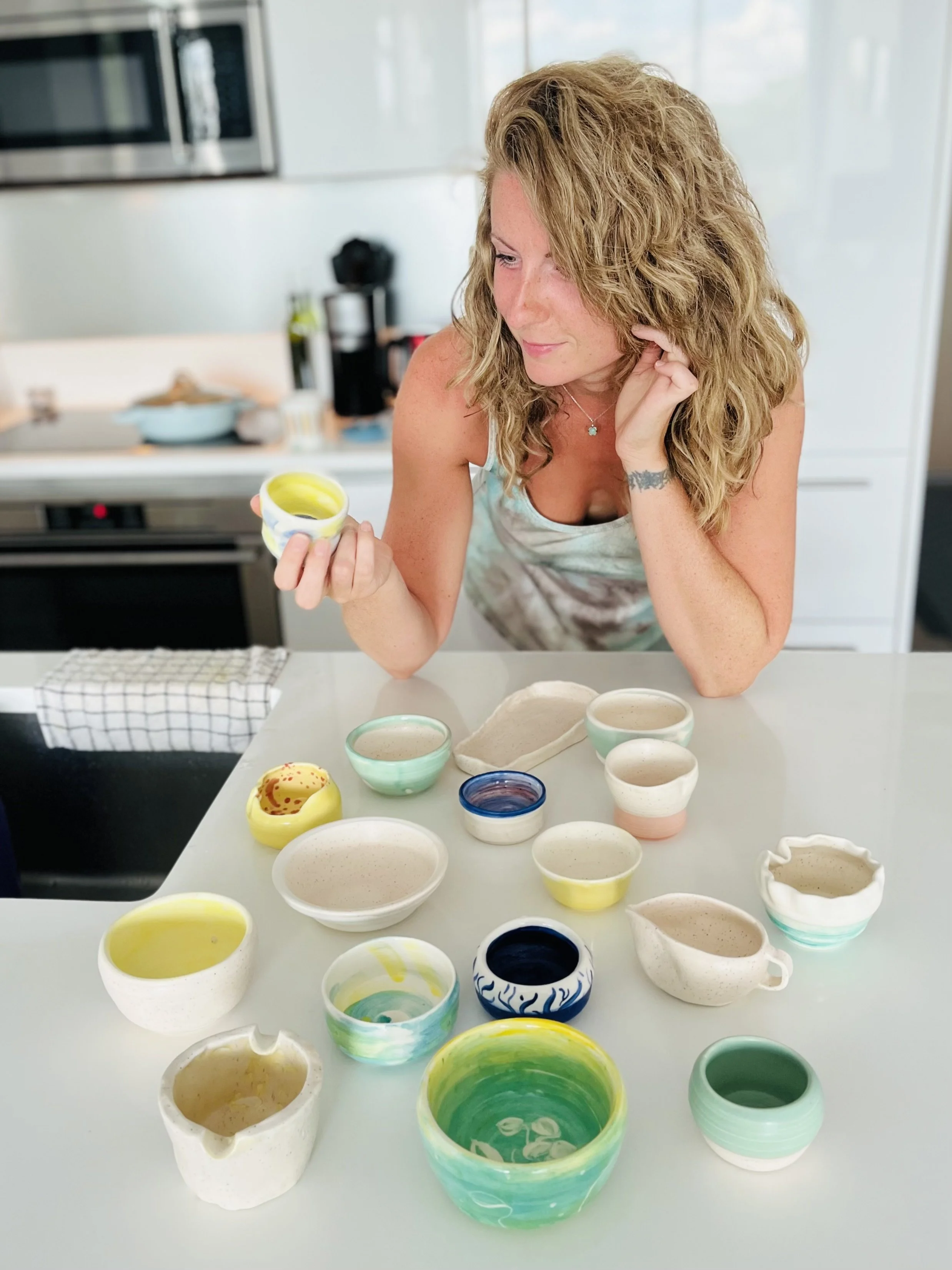 Woman with curly blonde hair looking at small colorful ceramic bowls on a white table in a modern kitchen.