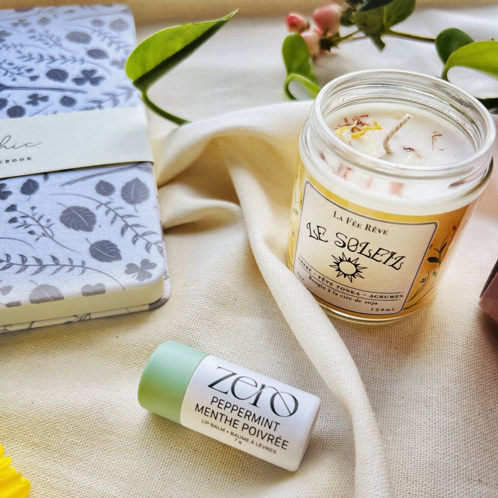 A desk setup with a notebook, a lit candle in a glass jar labeled "Les Solzets," a peppermint lip balm, a green leaf, pink flowers, and a white cloth on a beige fabric surface.
