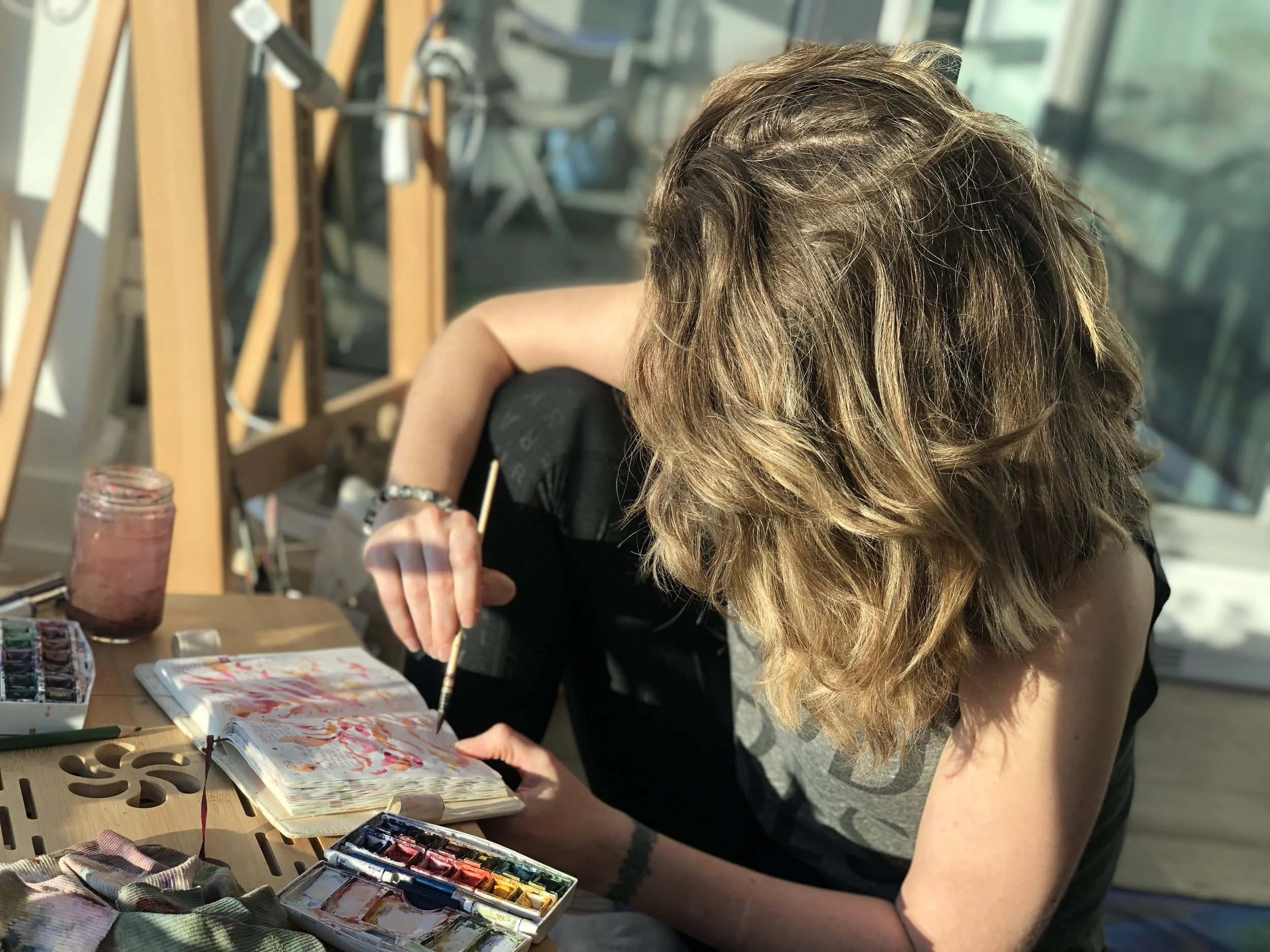 A person with long, curly, light brown hair is sitting at a table, painting in a sketchbook using watercolor paints. The table is scattered with painting supplies, including a palette, brushes, and fabric. The scene is well-lit by natural light coming through large windows.