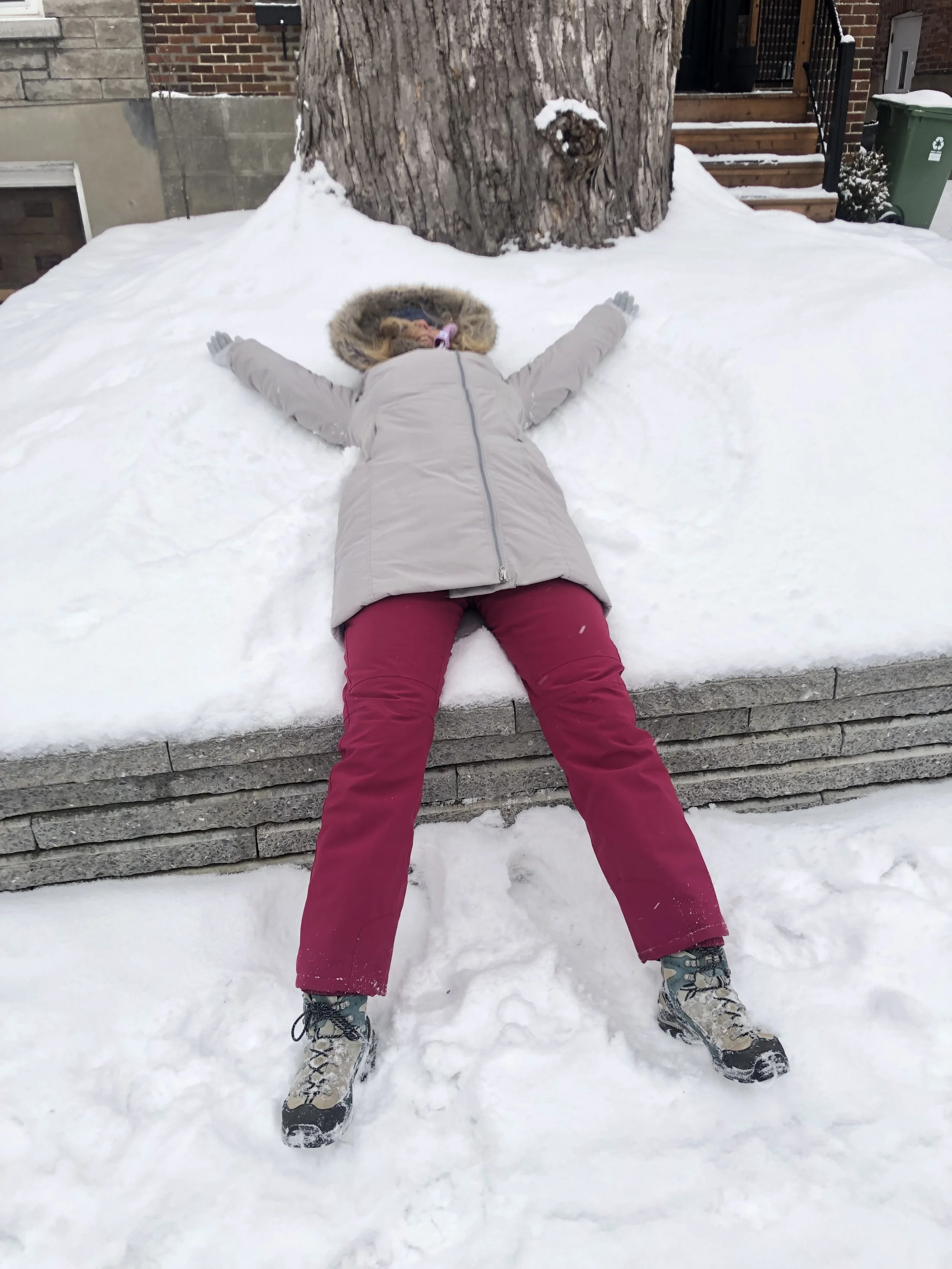 Person lying on their back in the snow, making a snow angel, wearing a beige winter coat with a fur-lined hood, red pants, and snow boots. There is a large tree and a brick building with stairs and a green recycling bin in the background.