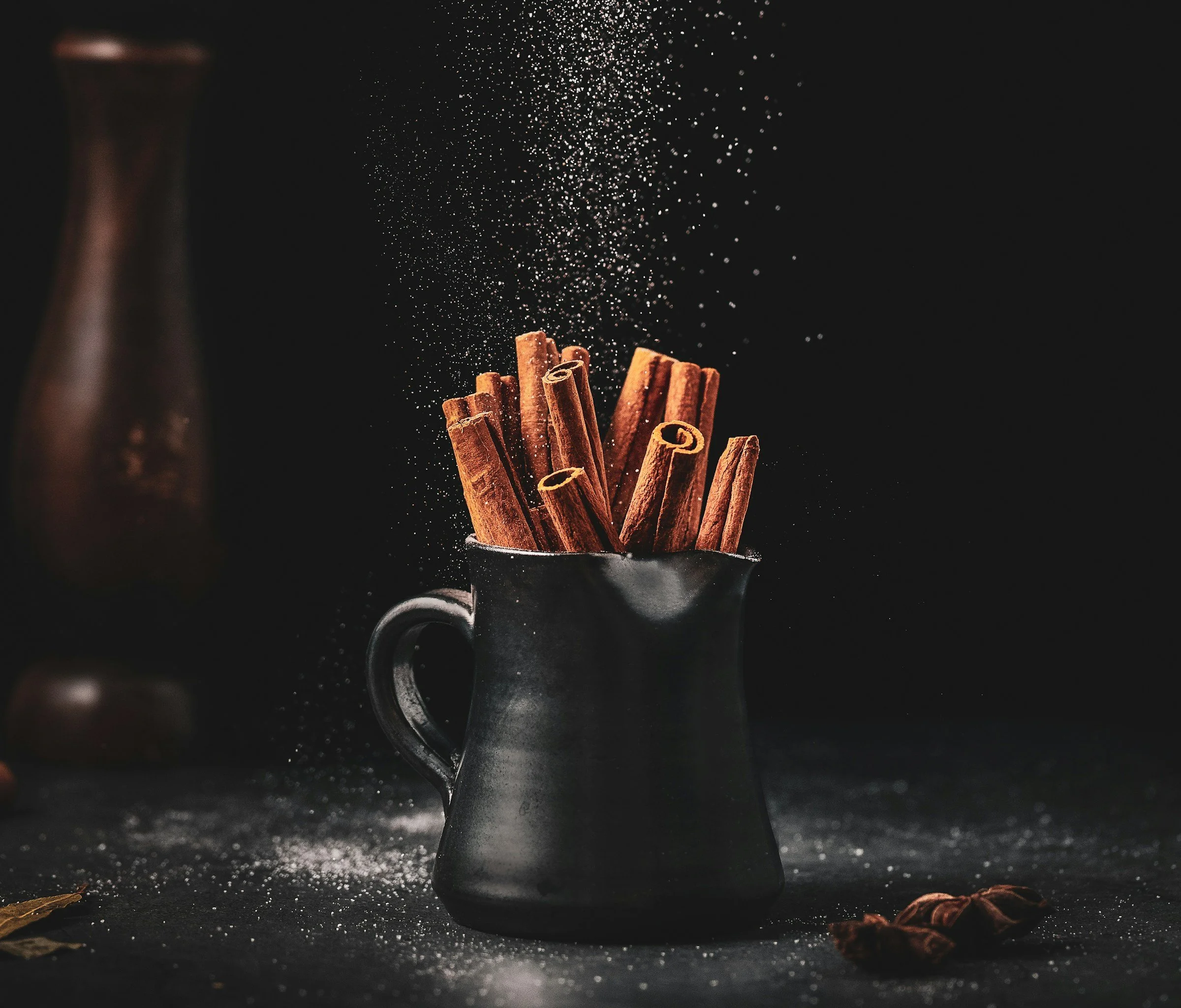 Cinnamon sticks inside a dark gray mug with a handle, with cinnamon powder and whole cinnamon sticks scattered on a black surface, and falling cinnamon powder creating a cloud above.