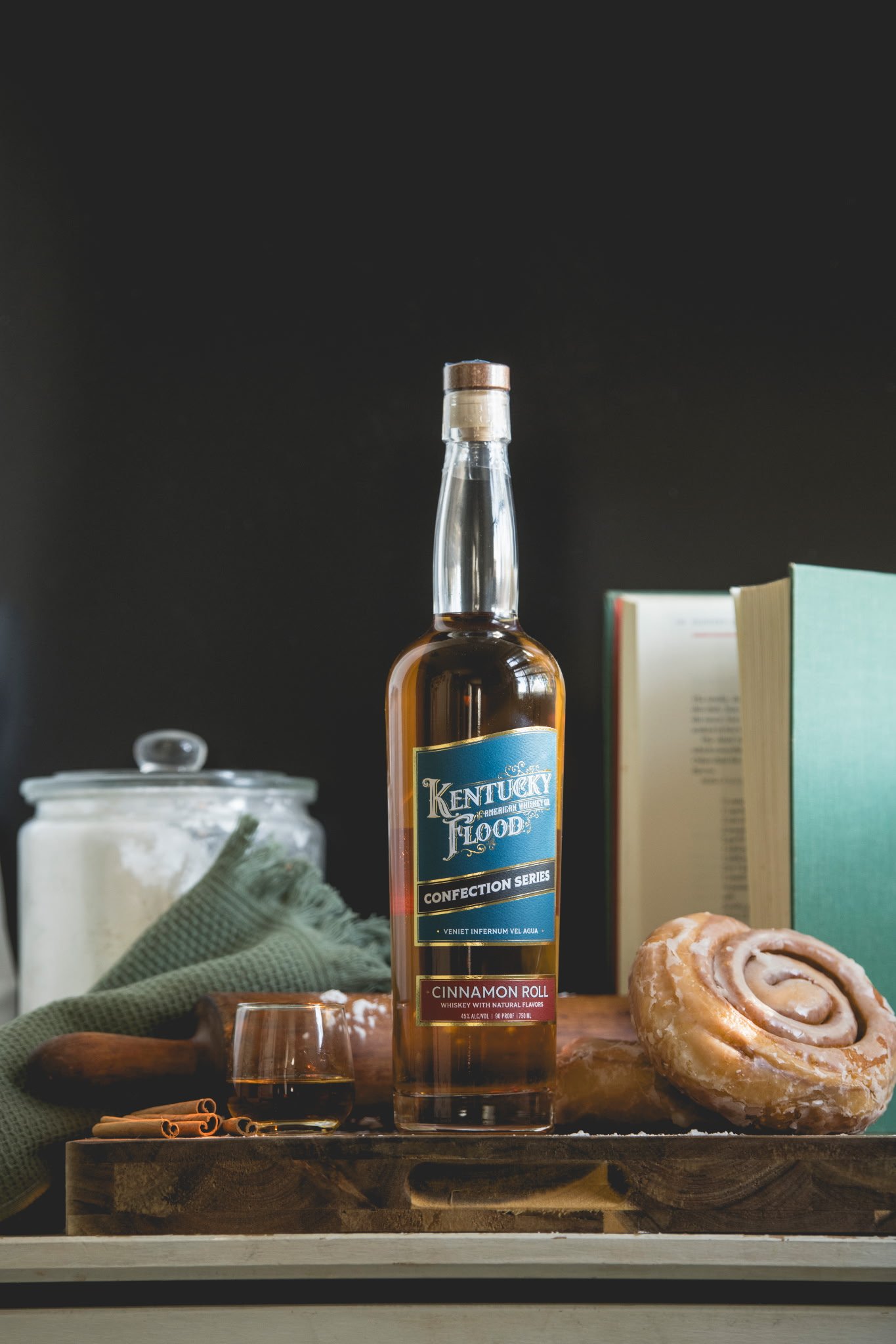 A bottle of Kentucky Flood whiskey on a wooden tray, with cinnamon rolls and cinnamon sticks, green cloth, glassware, a jar, and open books in the background against a black wall.