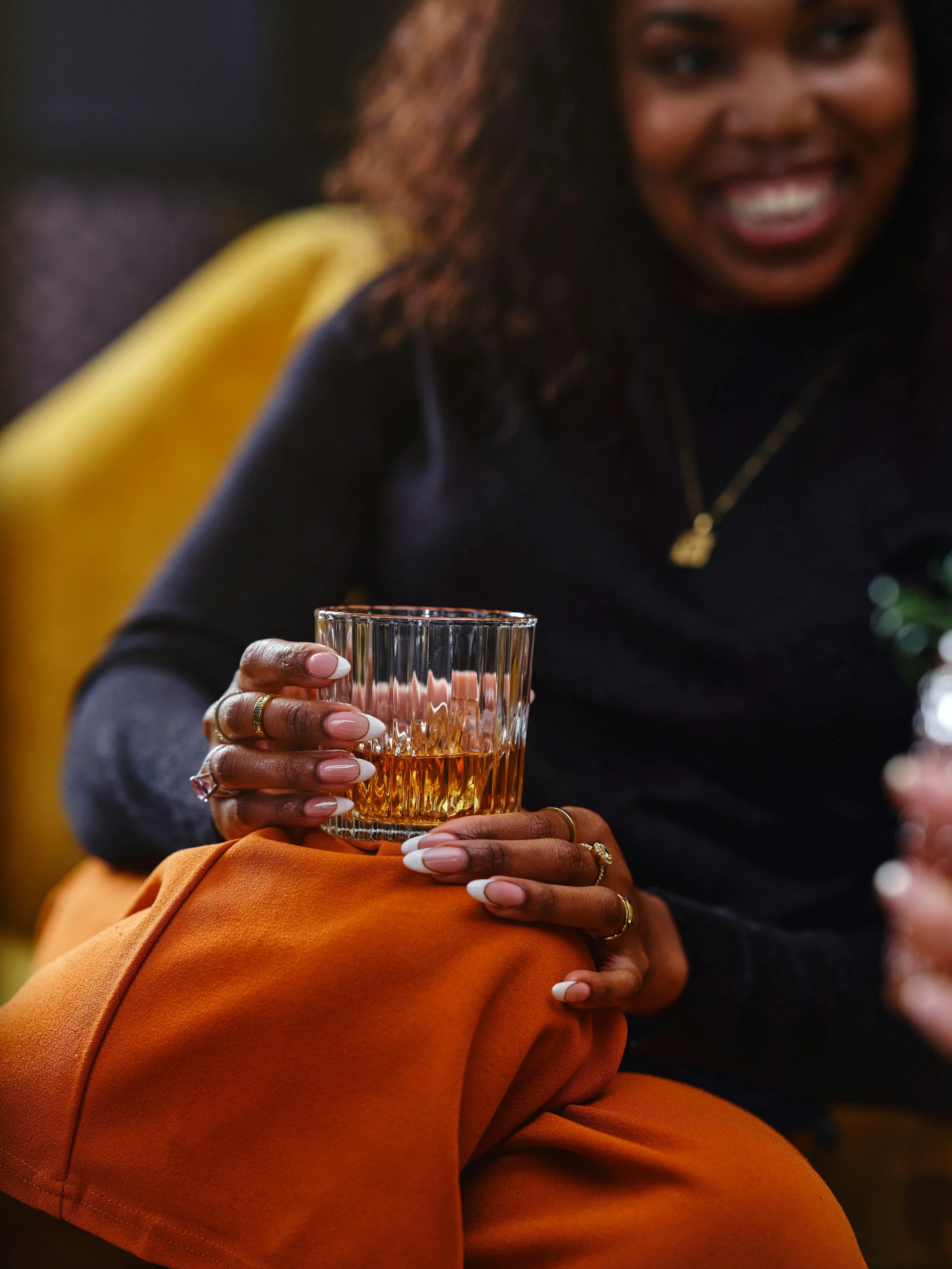 A woman with dark skin, wearing rings, with long nails, holding a glass of drink, sitting on a person wearing orange pants.