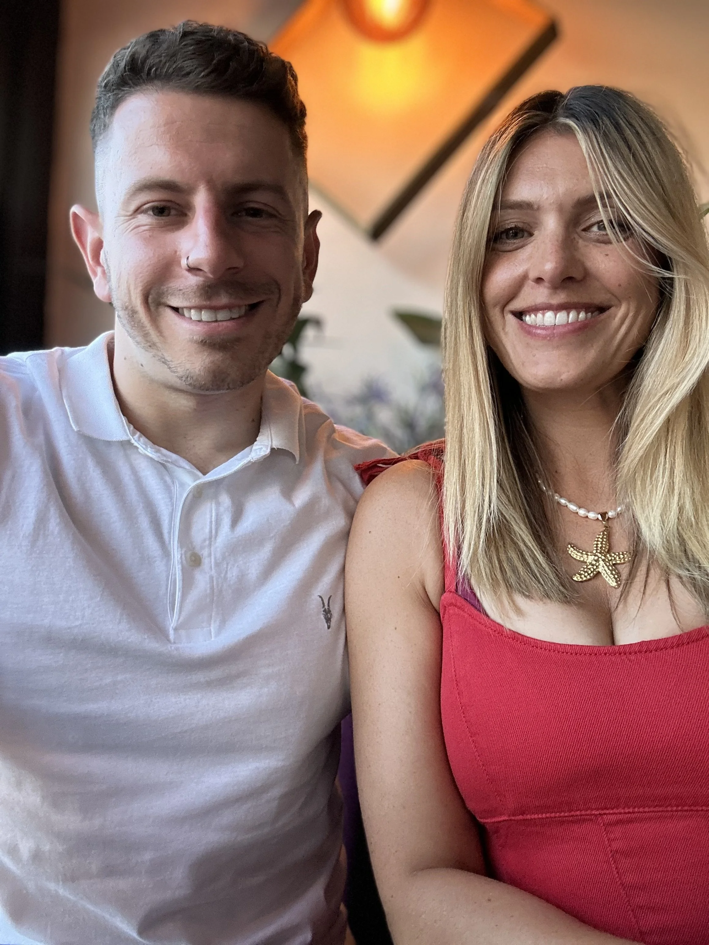 Close-up of a smiling man in a white shirt and a smiling woman in a red top and starfish necklace, sitting indoors with warm lighting.