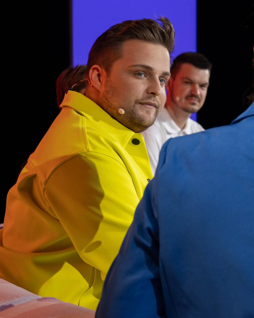 Drei Männer sitzen an einem Tisch bei einer Veranstaltung, der Mann im Vordergrund trägt eine gelbe Jacke.