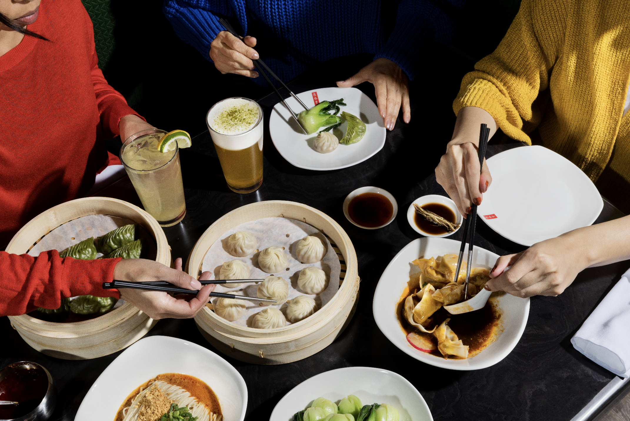 Image of a Taiwanese dinner spread featuring xiao long bao, dumplings, bok choy, and various beverages. 3 women enjoying the food in the scene