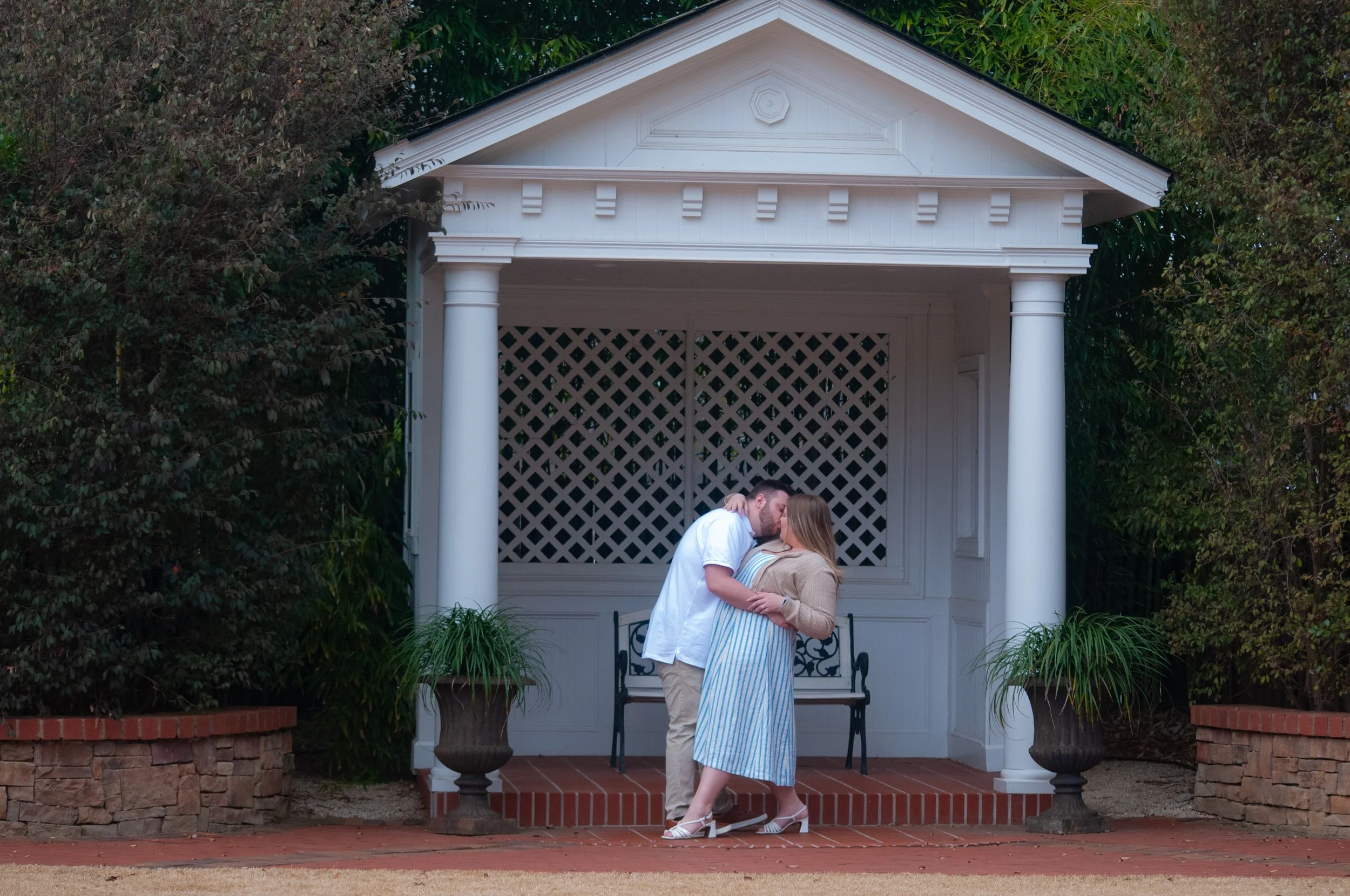 A couple sharing a romantic kiss in front of a white gazebo surrounded by greenery.