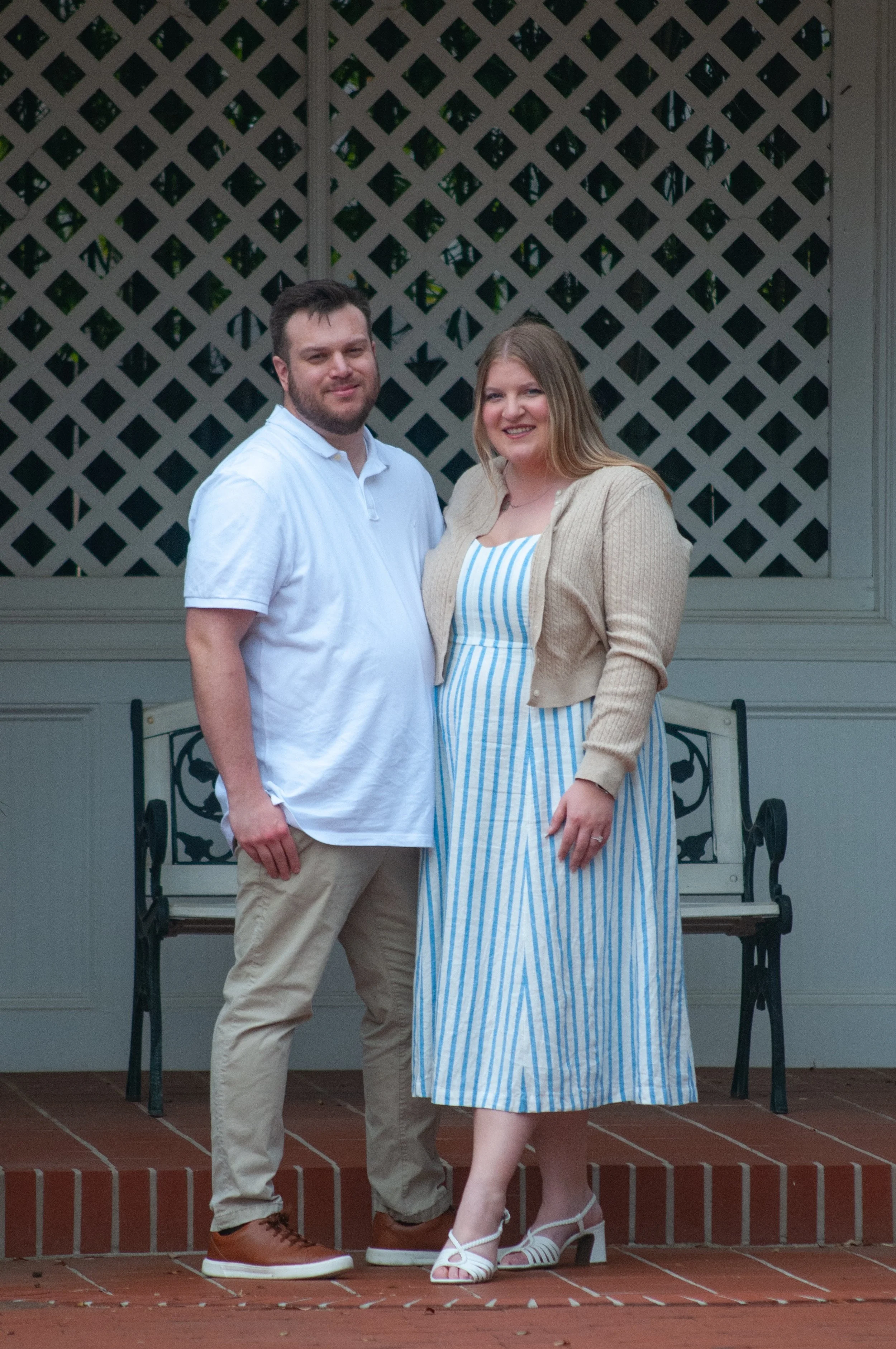 A young couple poses on a brick patio in front of a lattice screen and a white wall, dressed in casual summer clothing, smiling at the camera.