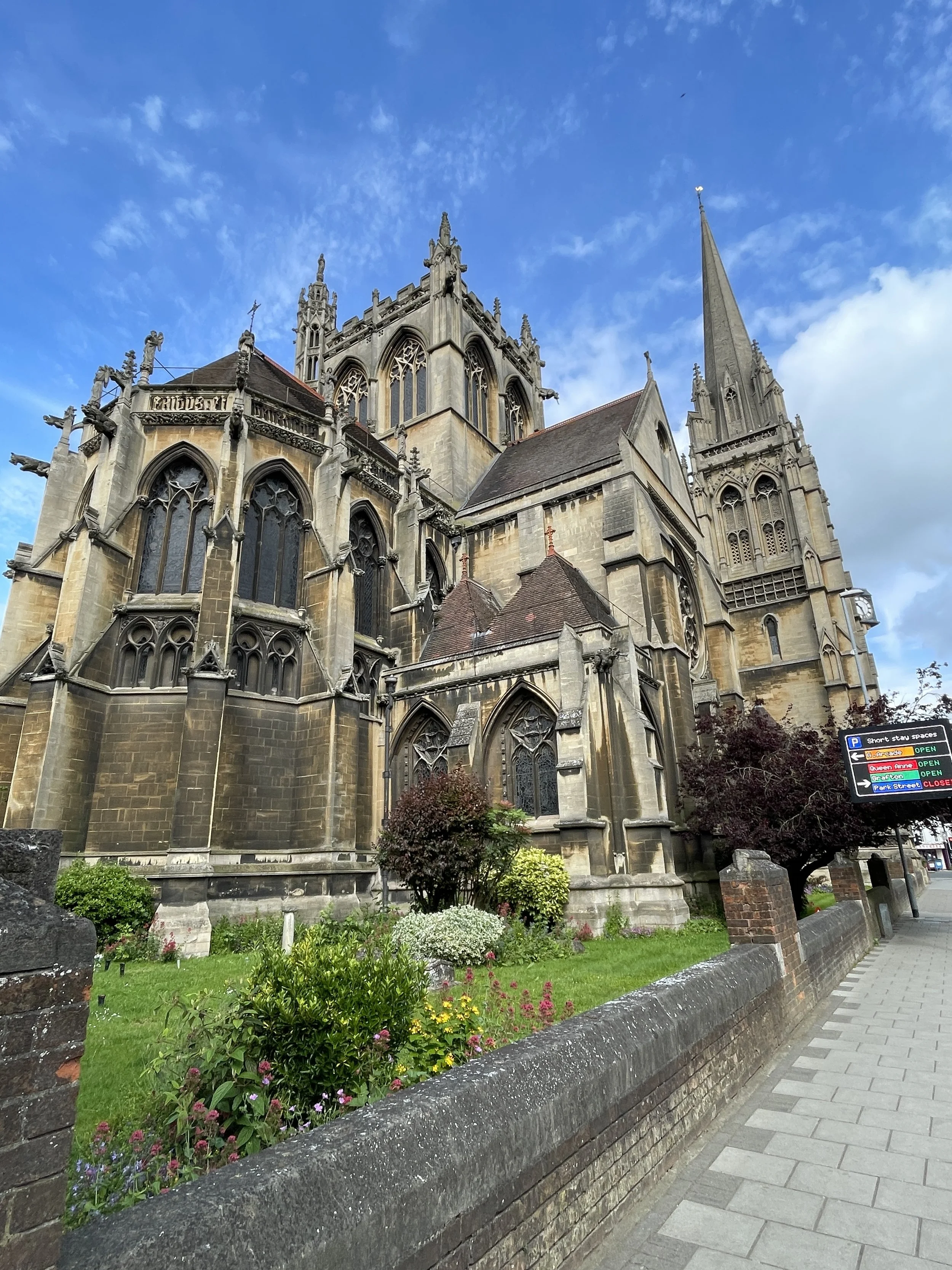 A large Gothic-style church with ornate stone carvings, tall stained glass windows, and spires reaching towards a partly cloudy blue sky. There is a landscaped area with flowers and bushes in front, and a sidewalk with a brick wall along the street.