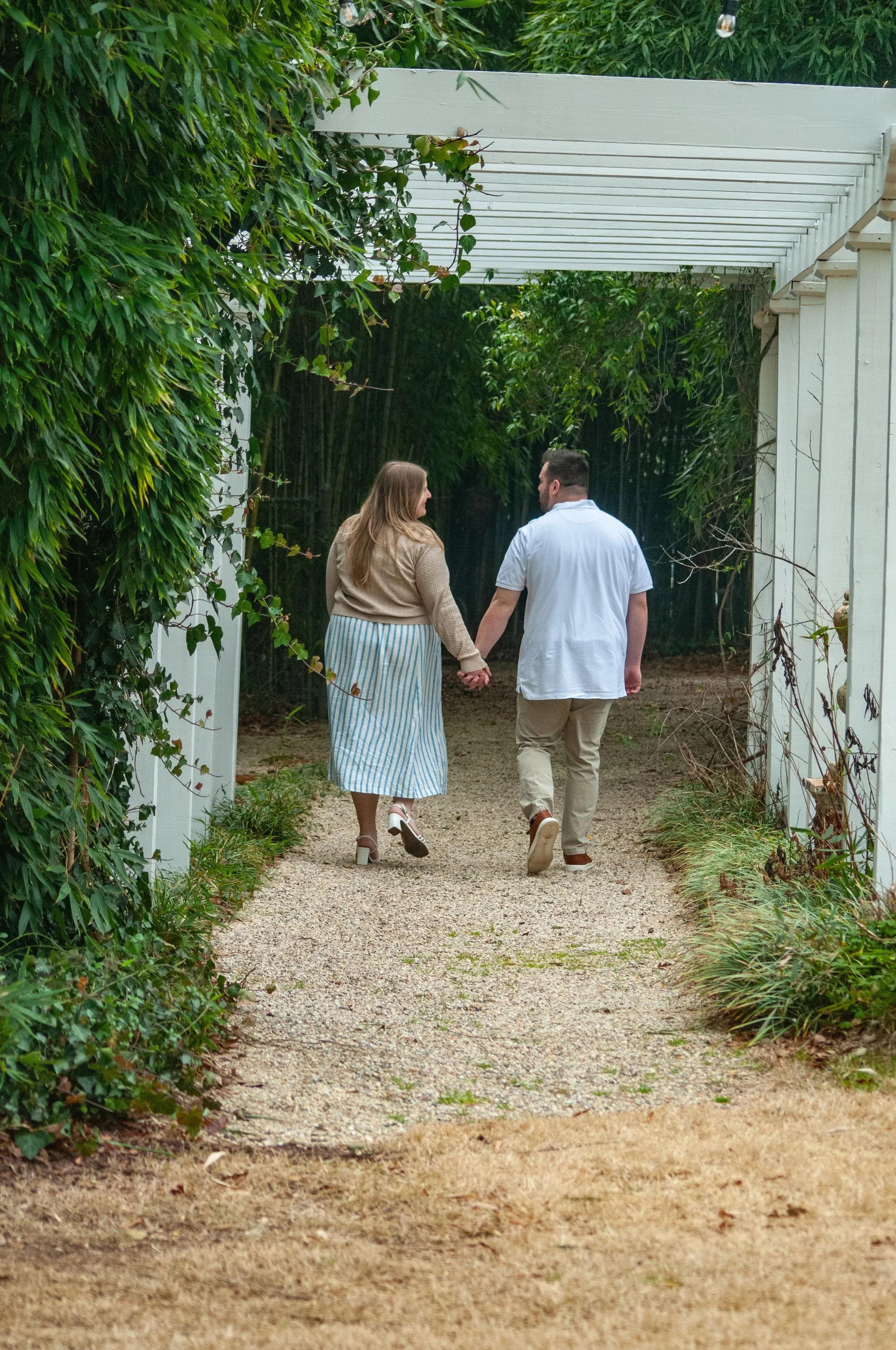 A couple walking hand in hand through a green, leafy garden pathway, with shrubs on both sides and a white trellis overhead.