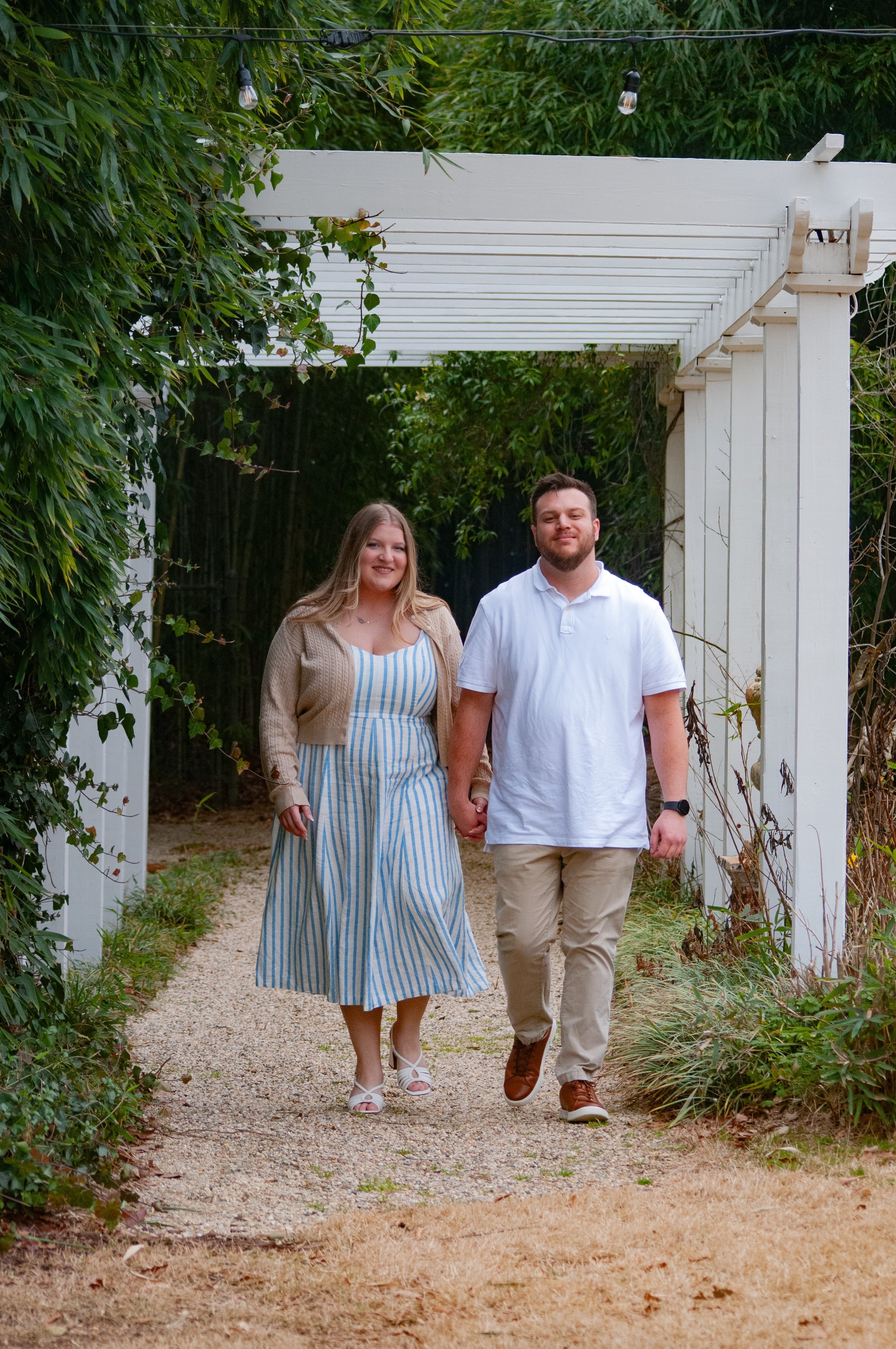 A couple walking hand in hand through a garden archway, with greenery and string lights above them, smiling and dressed casually.