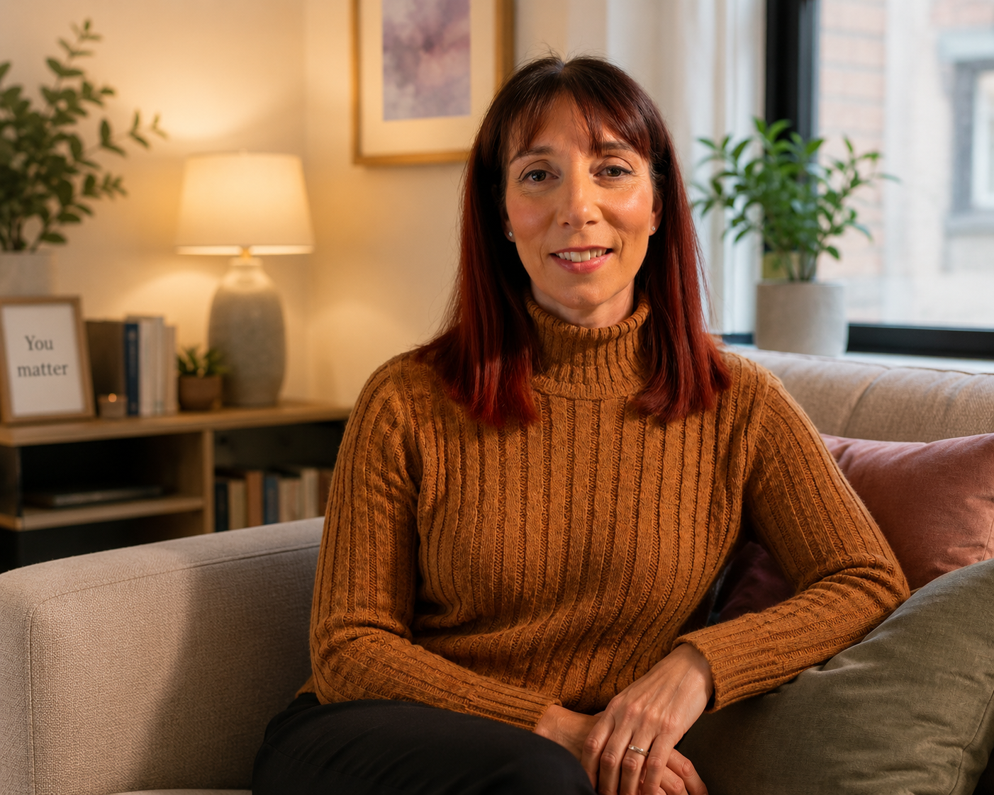 A woman with reddish-brown hair wearing an orange turtleneck sweater sitting on a beige sofa in a cozy living room with a lamp, plants, and framed art on the wall.