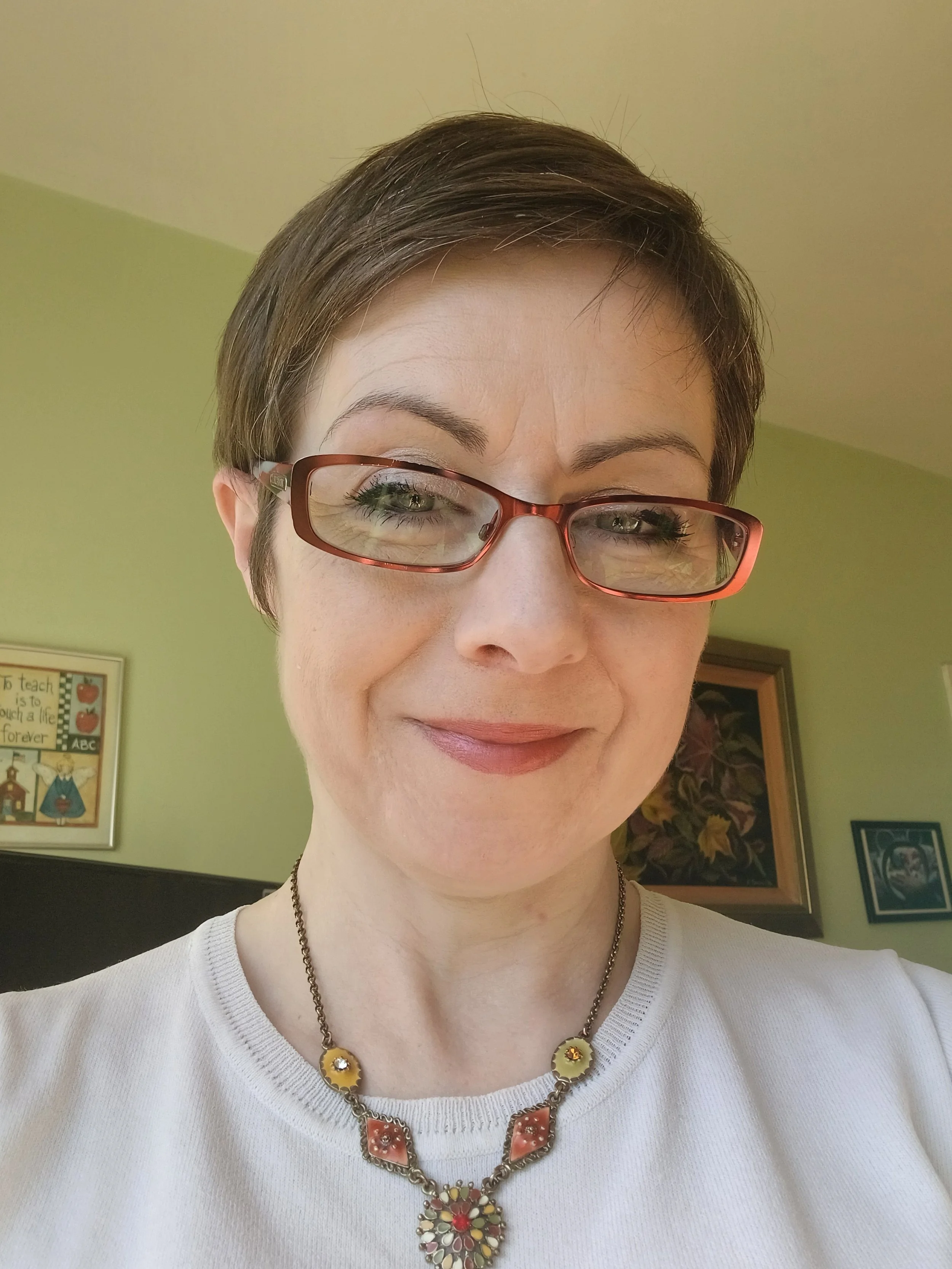 A woman with short brown hair, glasses with red frames, and wearing a white top and colorful jewelry, smiling indoors with framed artwork on the wall behind her.