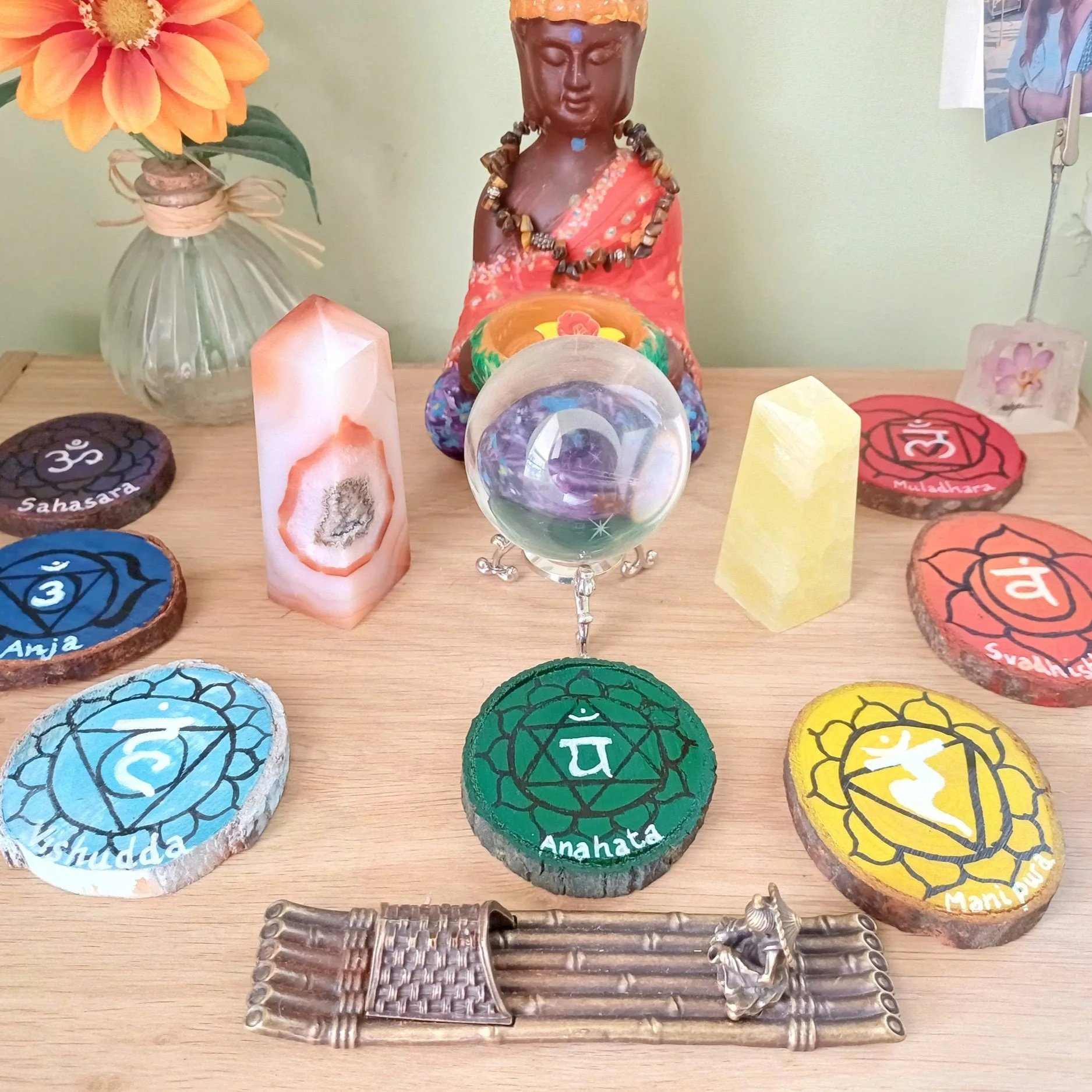 Set of painted rocks with chakra symbols and names, a brass wind chime, a candle, and a decorative Buddha figure with a necklace in the background.