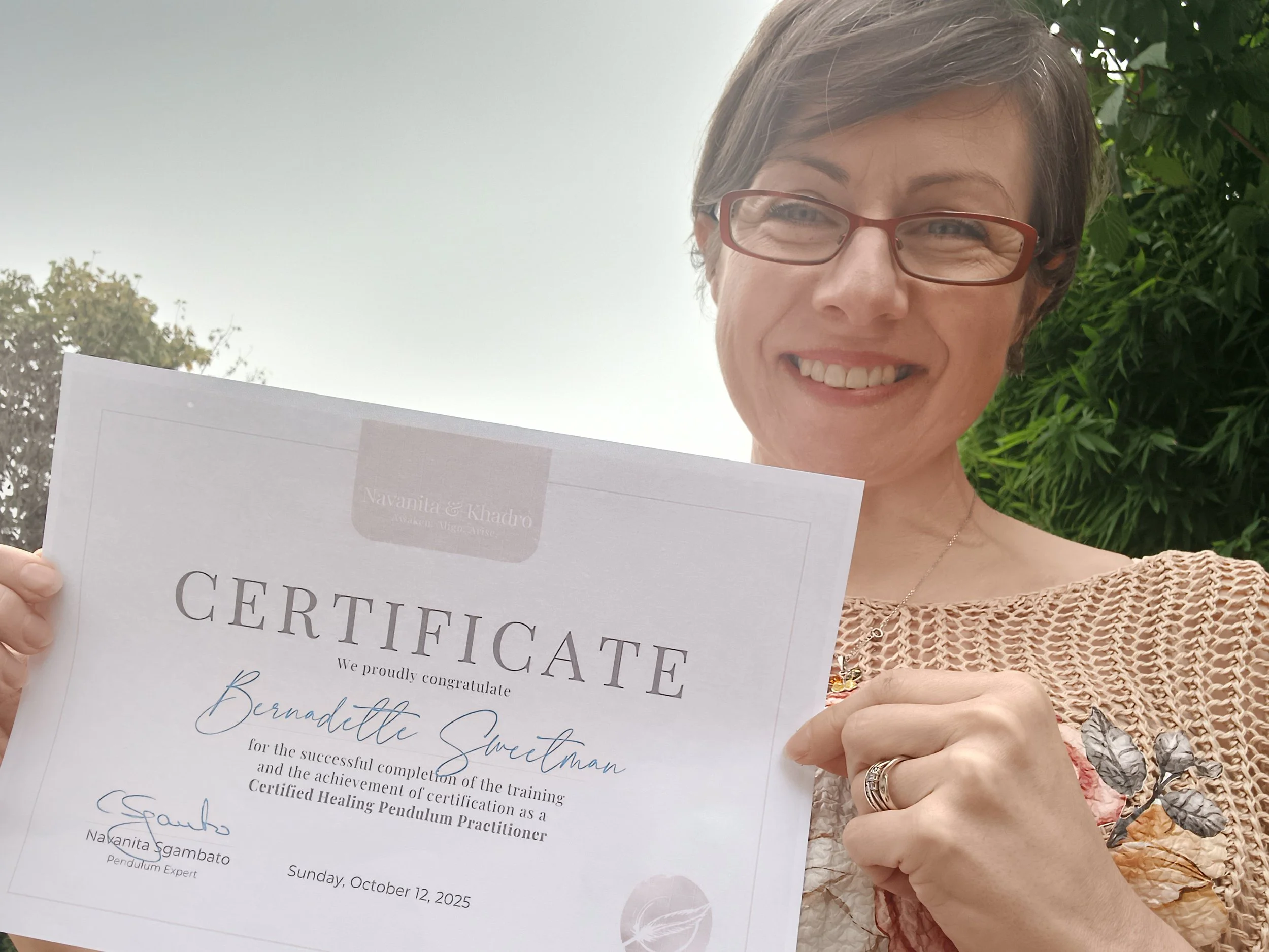 A woman with short brown hair, glasses, and a textured pink top is smiling while holding a certificate. She is outside, with green leafy bushes behind her, and a mostly gray sky overhead.