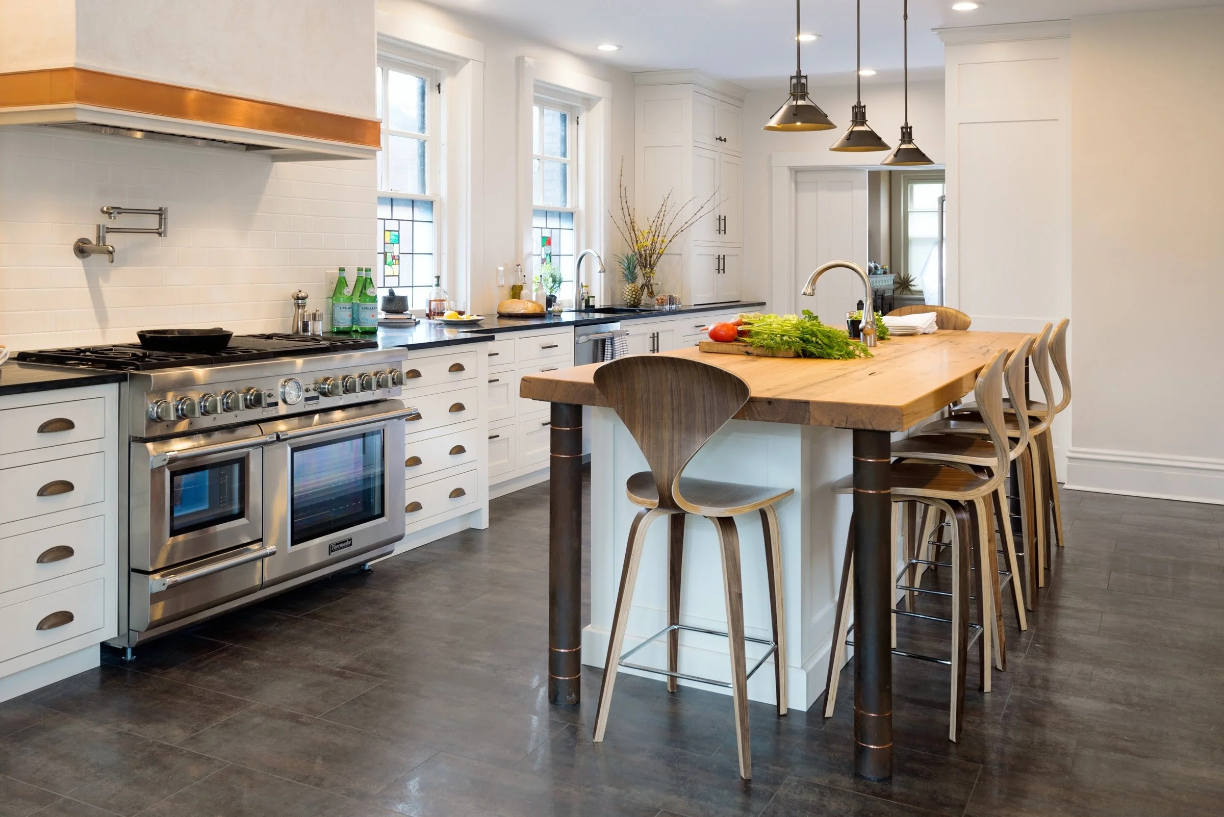 A modern kitchen with white cabinets, black countertops, a stainless steel stove, and a wooden island with multiple bar stools. There are three windows with stained glass accents, pendant lighting, and various plants and bottles on the counter.
