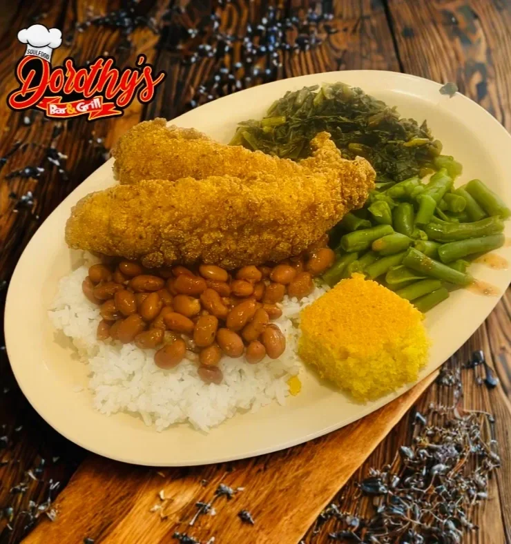 A plate of fried fish, white rice topped with baked beans, cooked greens, green beans, and a cornbread muffin on a wooden surface with a logo for Dorothy's Bar & Grill.