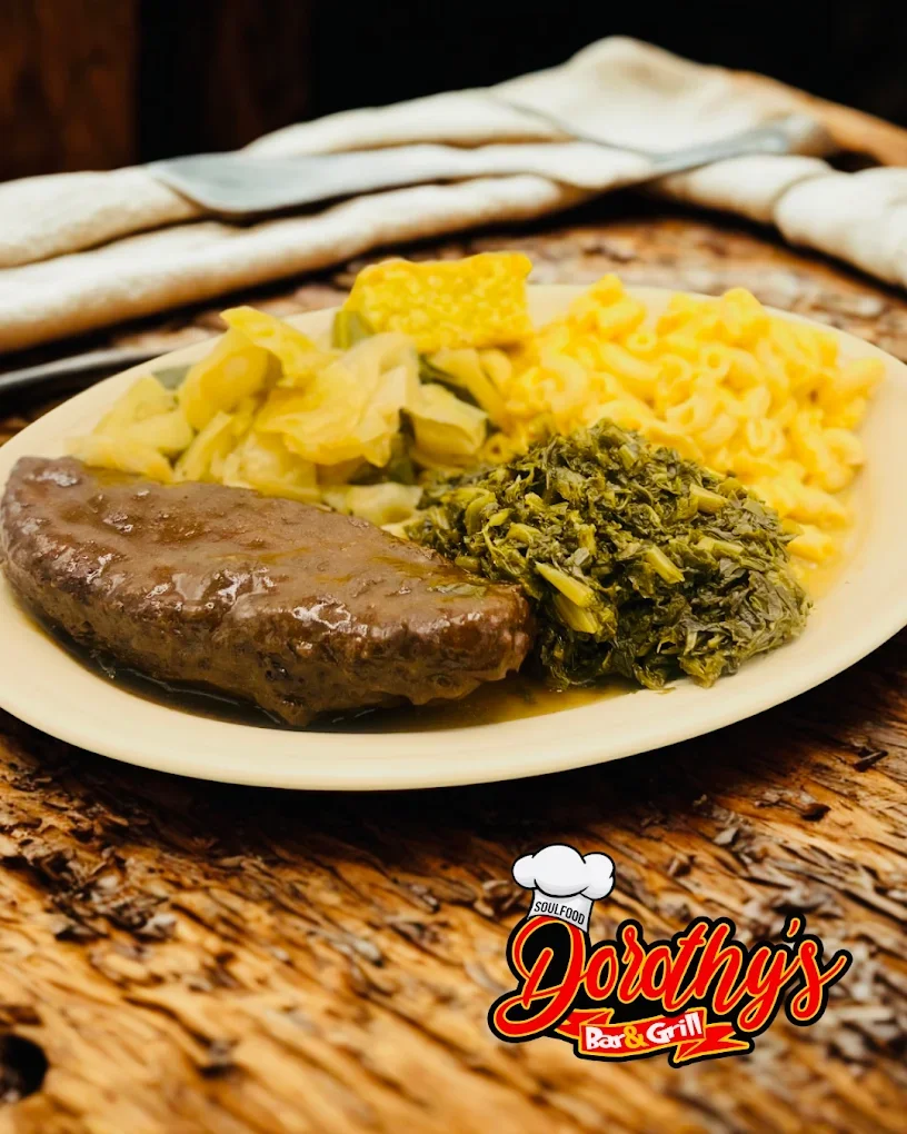 A plate of traditional Southern food including a grilled meatloaf, collard greens, mustard greens, macaroni and cheese, and a side of pickled yellow vegetables, served on a rustic wooden table. Dothy's Bar & Grill logo is at the bottom corner.