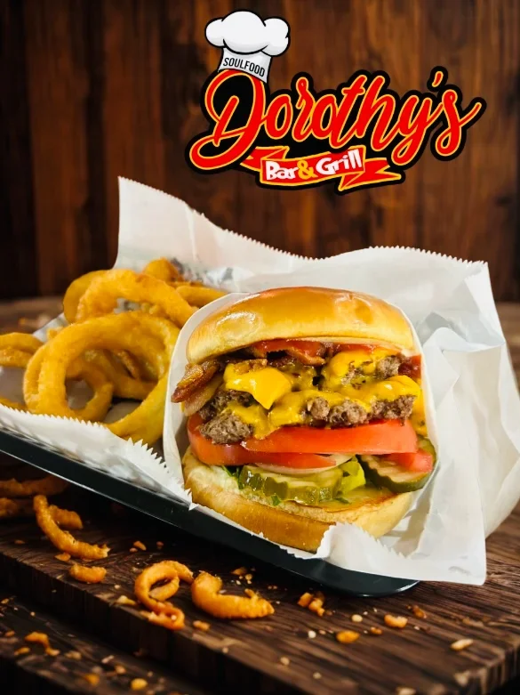 A cheeseburger with lettuce, tomato, pickles, onions, cheese, and beef patty in a bun, served with onion rings in a basket at Dorothy's Bar & Grill.