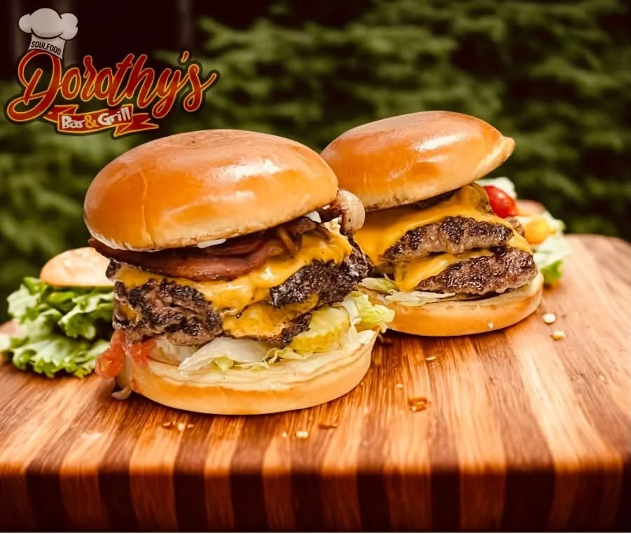 Two cheeseburgers with lettuce, tomato, pickles, onions, cheese, and beef patties on a wooden surface outdoors.