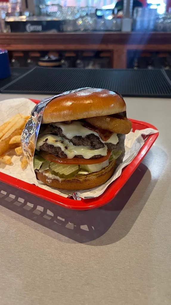 A burger with cheese, lettuce, tomato, pickles, onions, bacon, and onions rings in a bun, served on a red tray with fries.