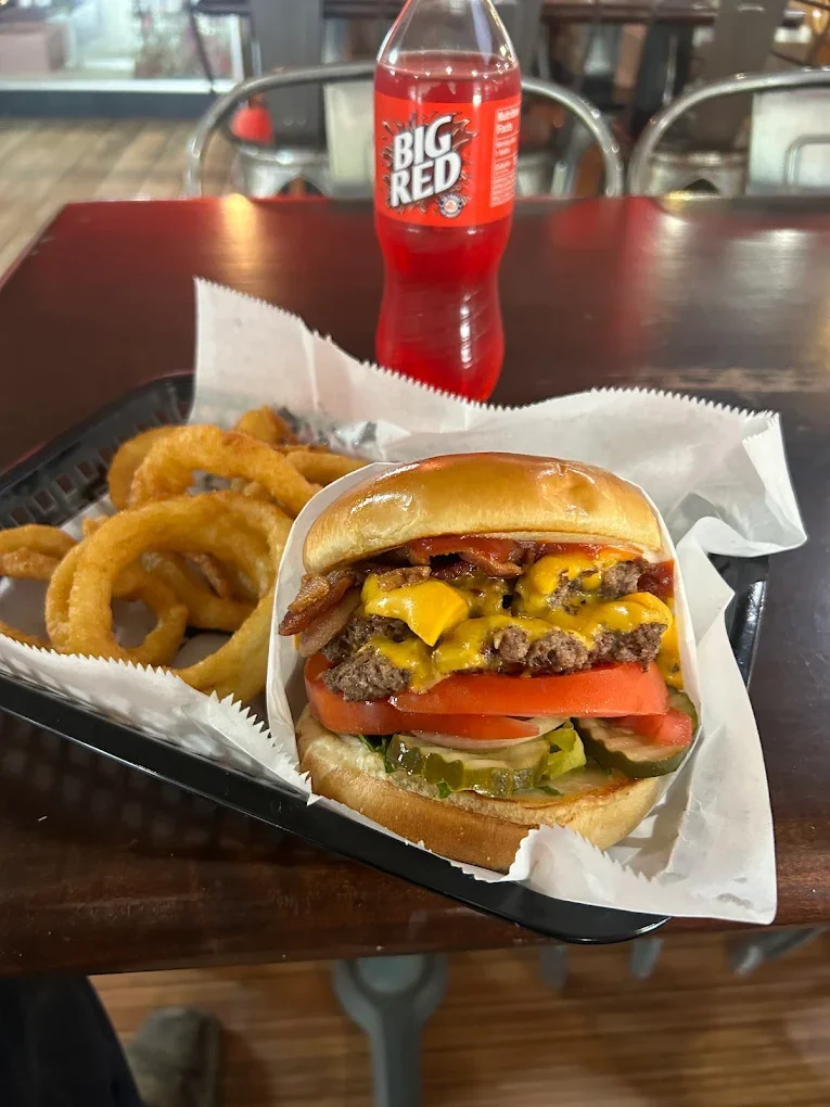 A cheeseburger with bacon, tomato, lettuce, pickles, and mustard on a bun, served with a side of onion rings, and a bottle of Big Red soda on a wooden table.