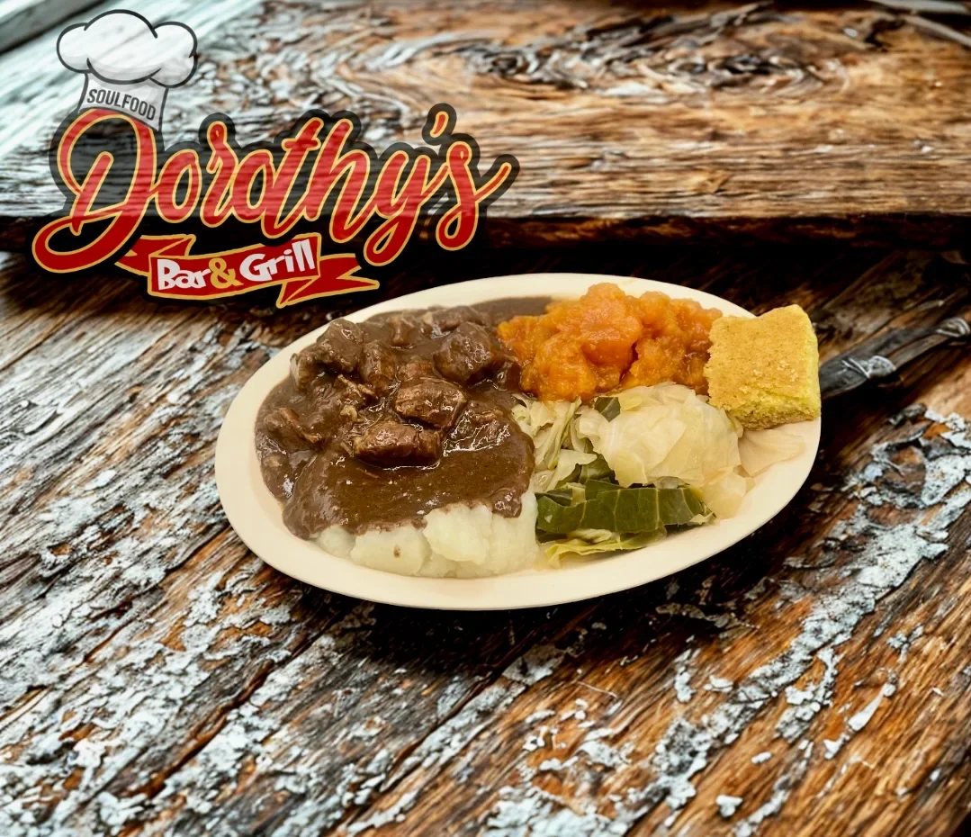 A plate of food with mashed potatoes, beef stew gravy, cooked vegetables, a scoop of orange mashed sweet potatoes, and a piece of cornbread, on a wooden table, with a sign that says 'Dorthy's Bar & Grill' and a chef hat illustration.