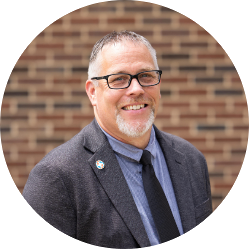 John C. Wilson, A smiling middle-aged man with glasses, a beard, and short gray hair, wearing a gray blazer, blue dress shirt, and black tie, standing in front of a brick wall.
