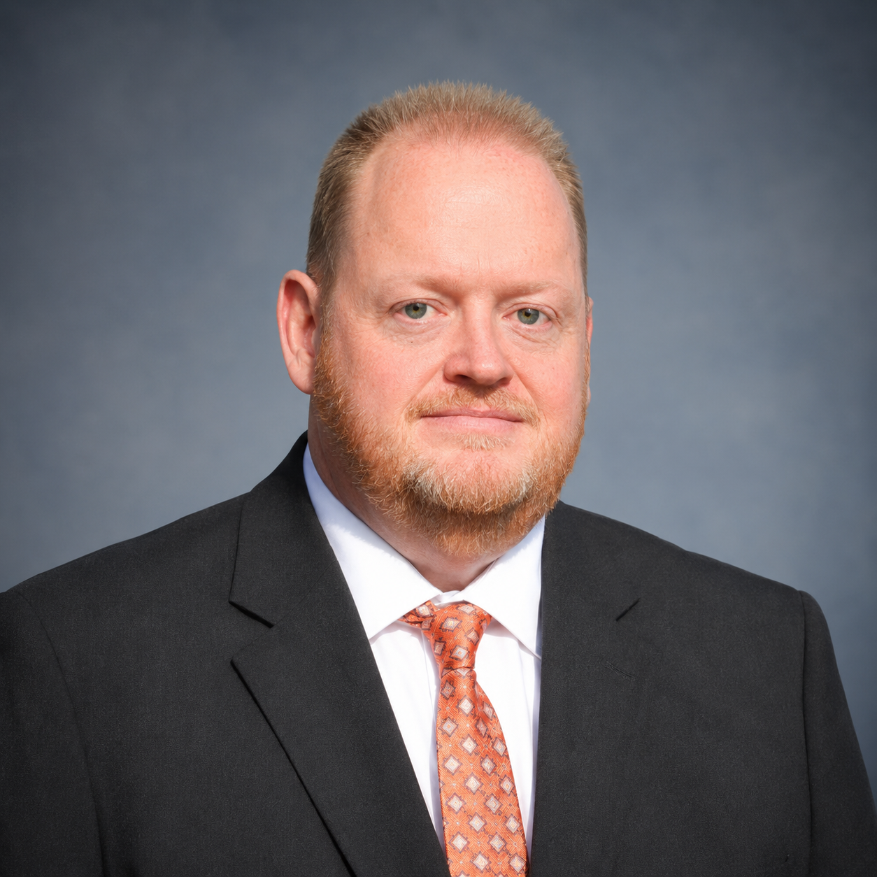 A professional headshot of a middle-aged man with light skin, reddish-blonde hair, and a beard, wearing a black suit, white shirt, and orange patterned tie against a neutral gray background.