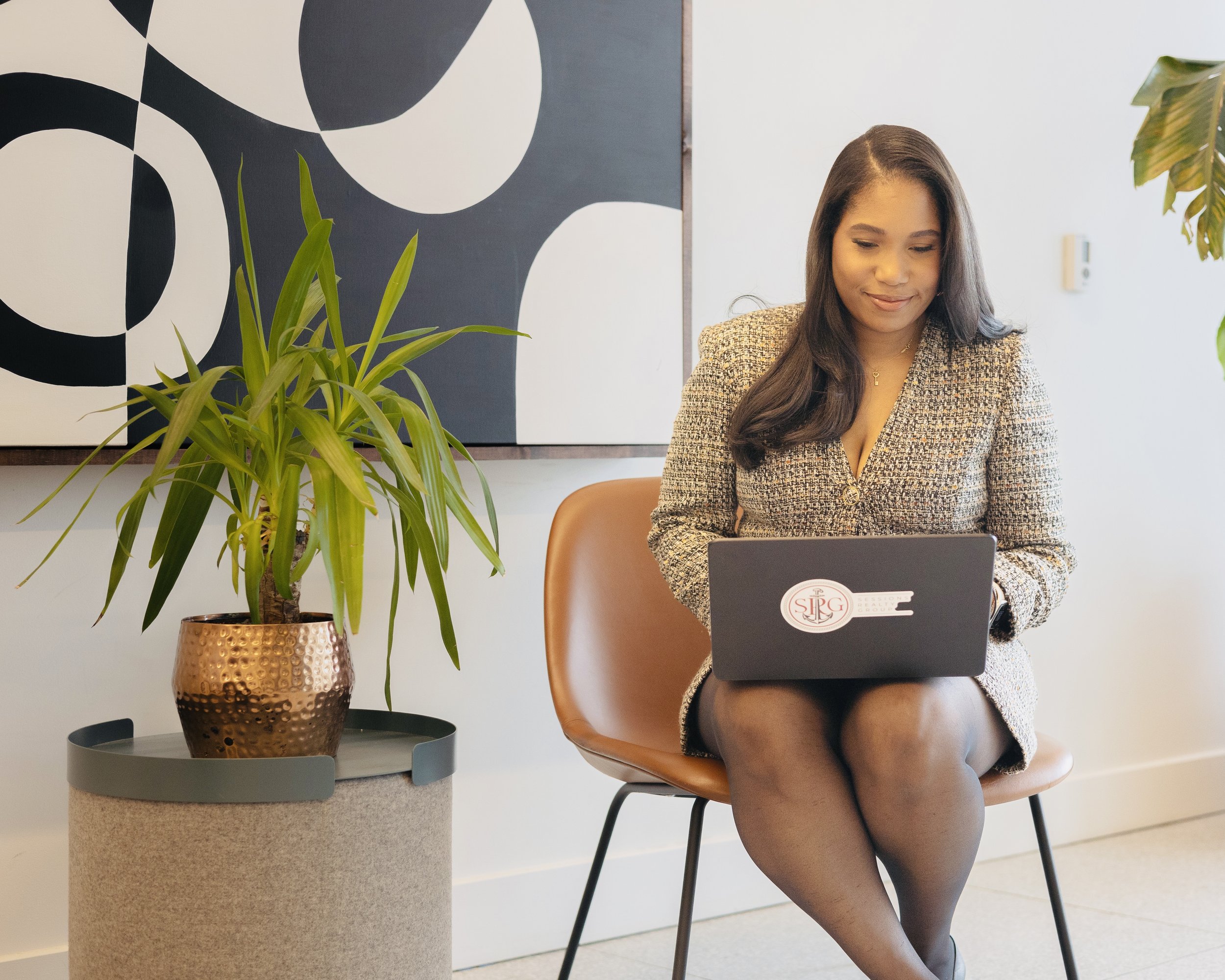 A woman sitting on a brown chair, dressed in a professional blazer, using a laptop, with a large green houseplant in a copper-colored pot on a side table nearby, and abstract black and white artwork on the wall behind her.