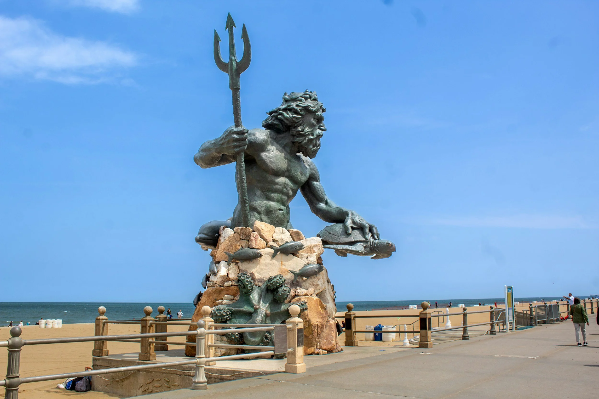 A large statue of a mythological sea god with a beard, holding a trident, sitting on rocks surrounded by marine life sculptures, near a beach with people and a blue sky in the background.