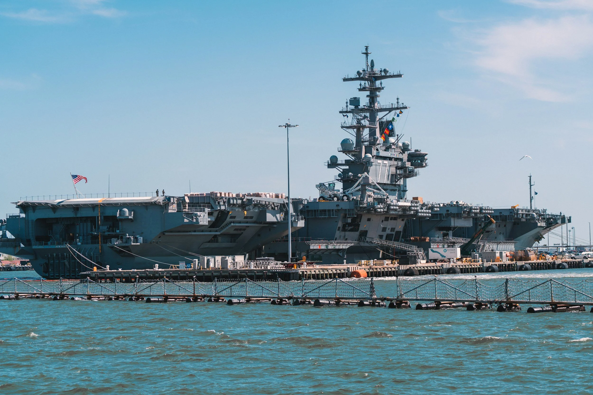 A large military aircraft carrier docked at a harbor with a clear sky in the background. The ship features various antennas, radar equipment, and a flag.