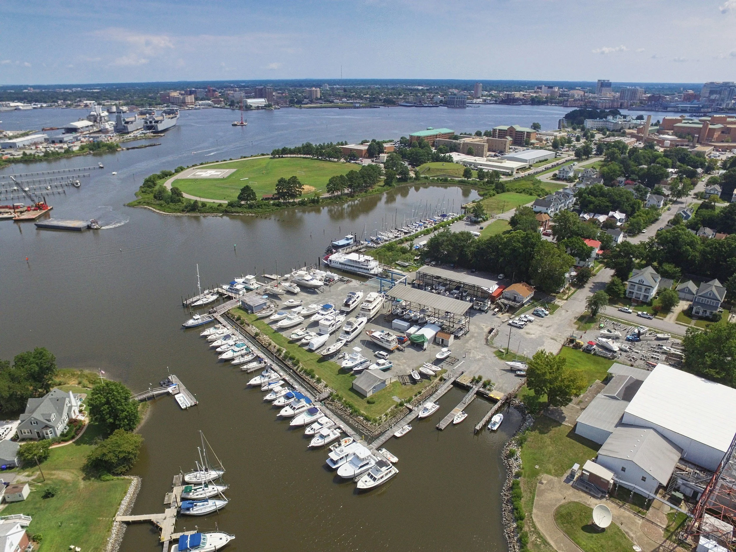 Aerial view of a harbor with numerous boats docked at piers, surrounded by residential houses, green parks, and a distant city skyline across a river under a partly cloudy sky.