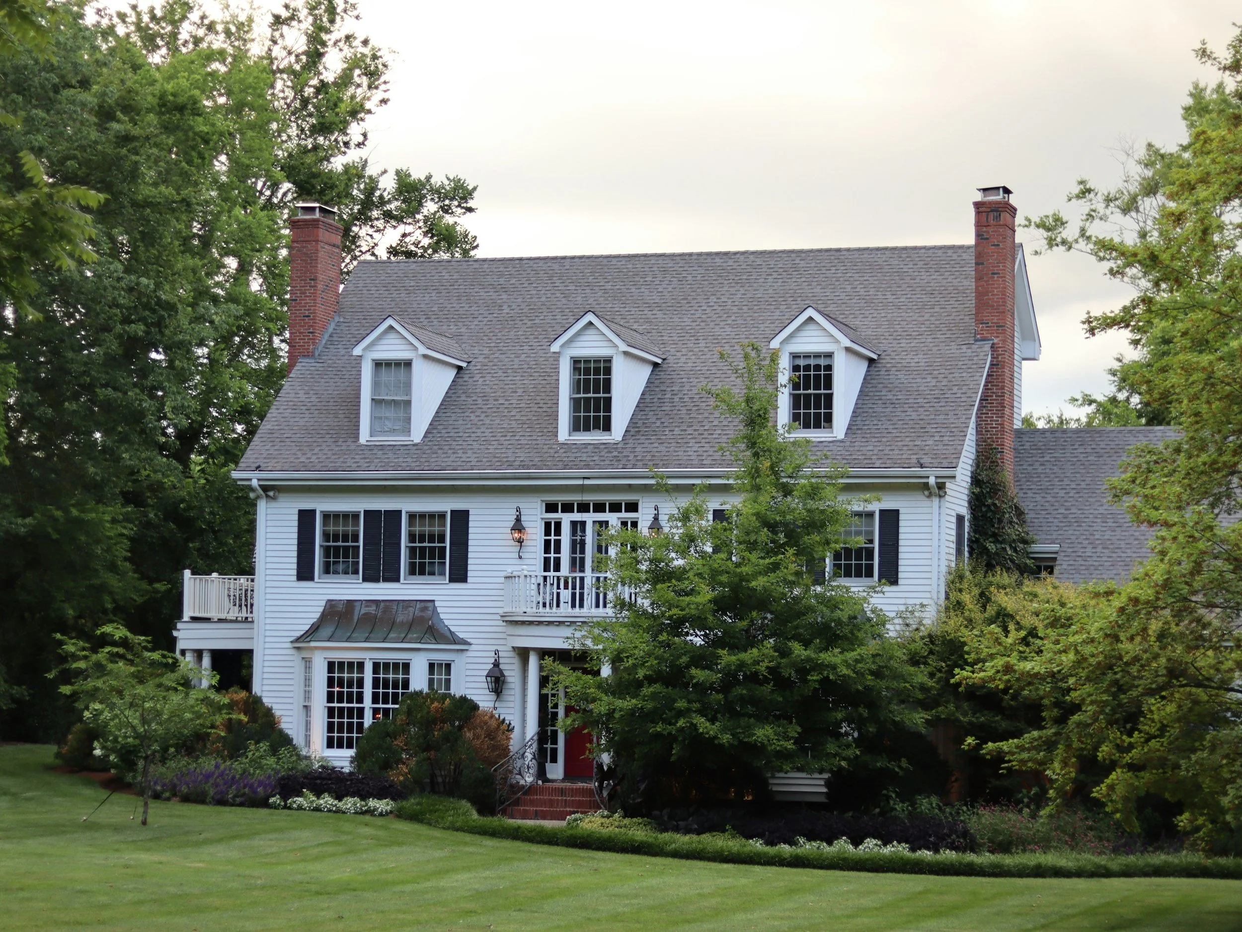 A large, white, two-story house with black shutters, multiple dormer windows, and brick chimneys, surrounded by green trees and a well-maintained lawn.