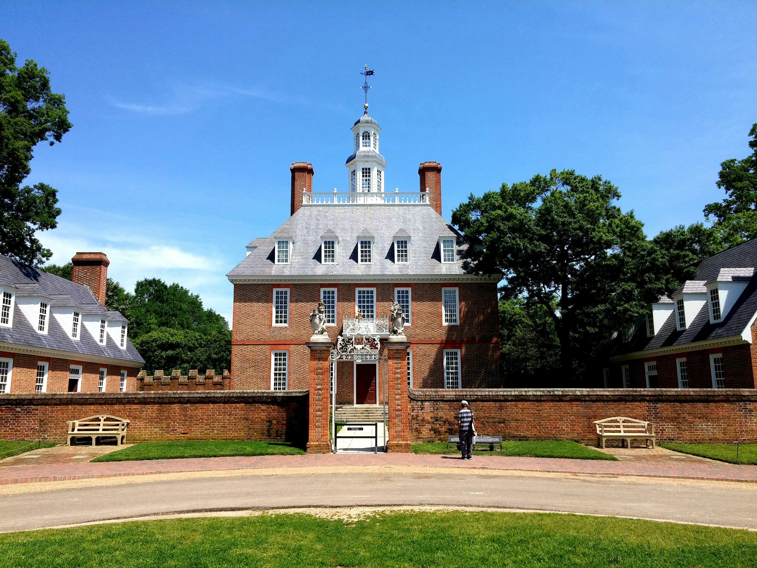 A historic brick building with a tower topped by a weather vane and a cupola, surrounded by trees and a brick wall, with benches and a person standing in front.